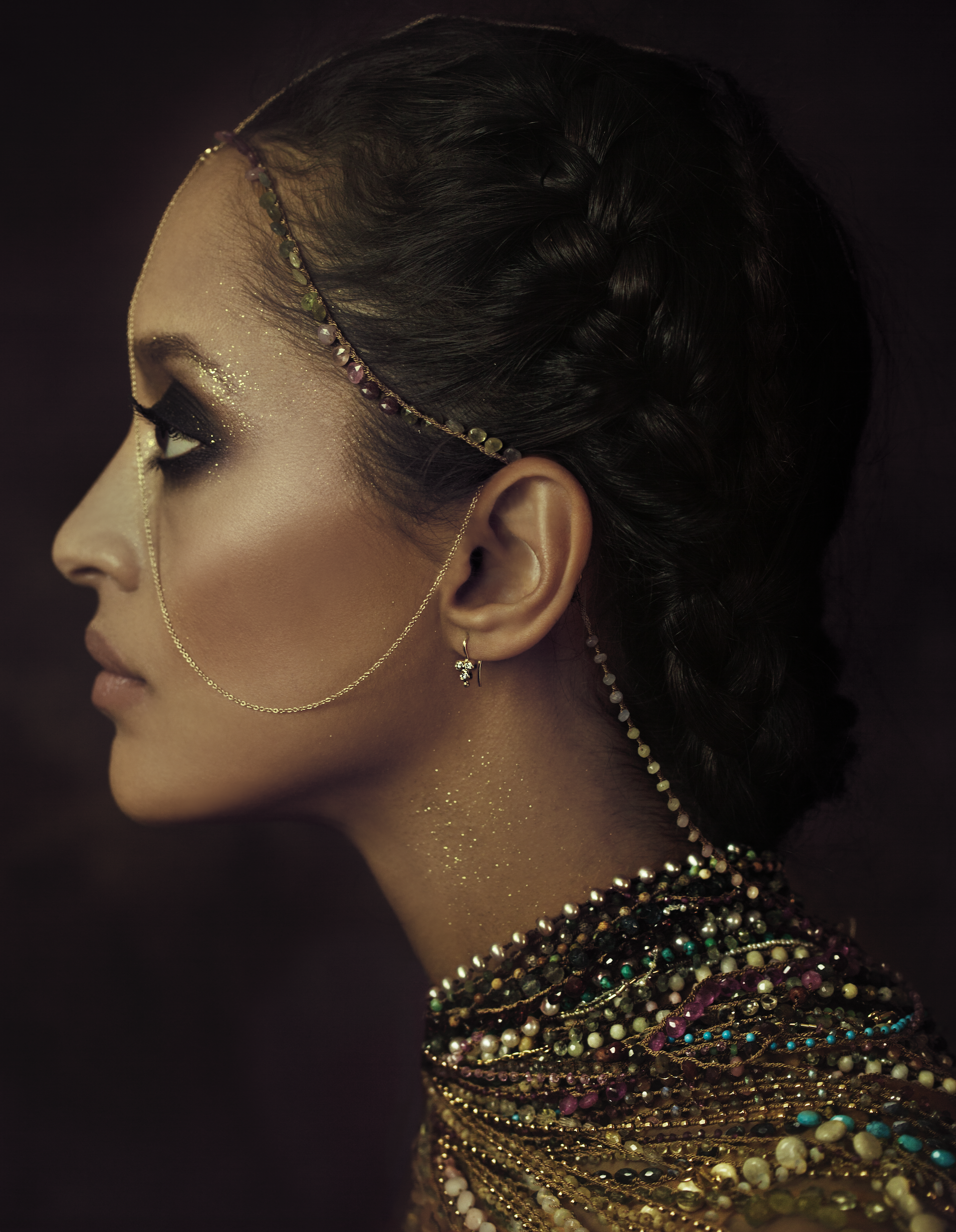 A woman with braided hair wearing intricate jewelry, including earrings, a delicate chain, and multiple beaded necklaces, with glitter makeup on her face and neck, set against a dark background.