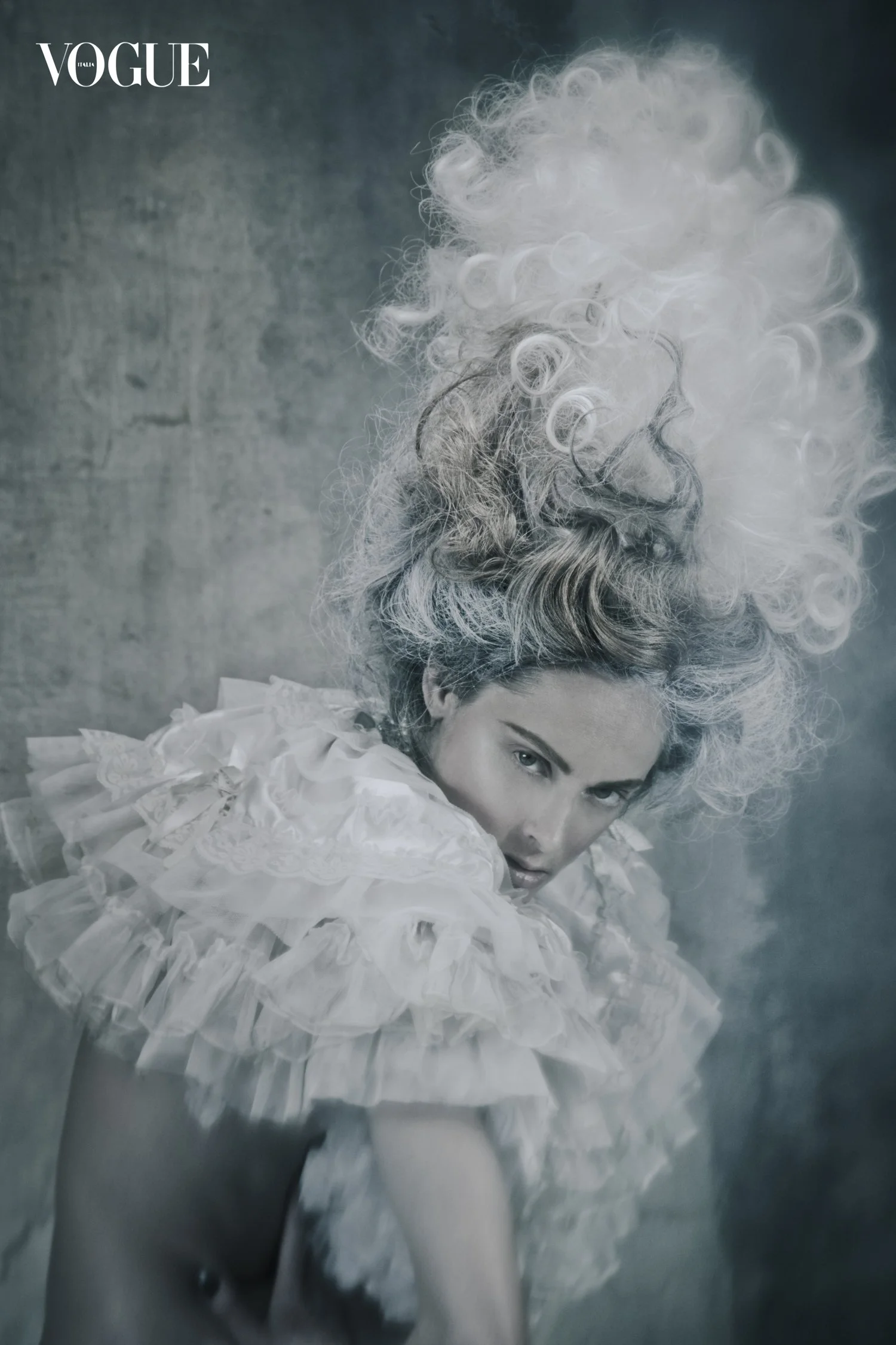 A woman dressed in a white frilly costume with a tall, curly, white and gray wig styled in an elaborate fashion, posing with her head tilted and looking intensely at the camera against a textured dark background. The Vogue logo is in the top left cor