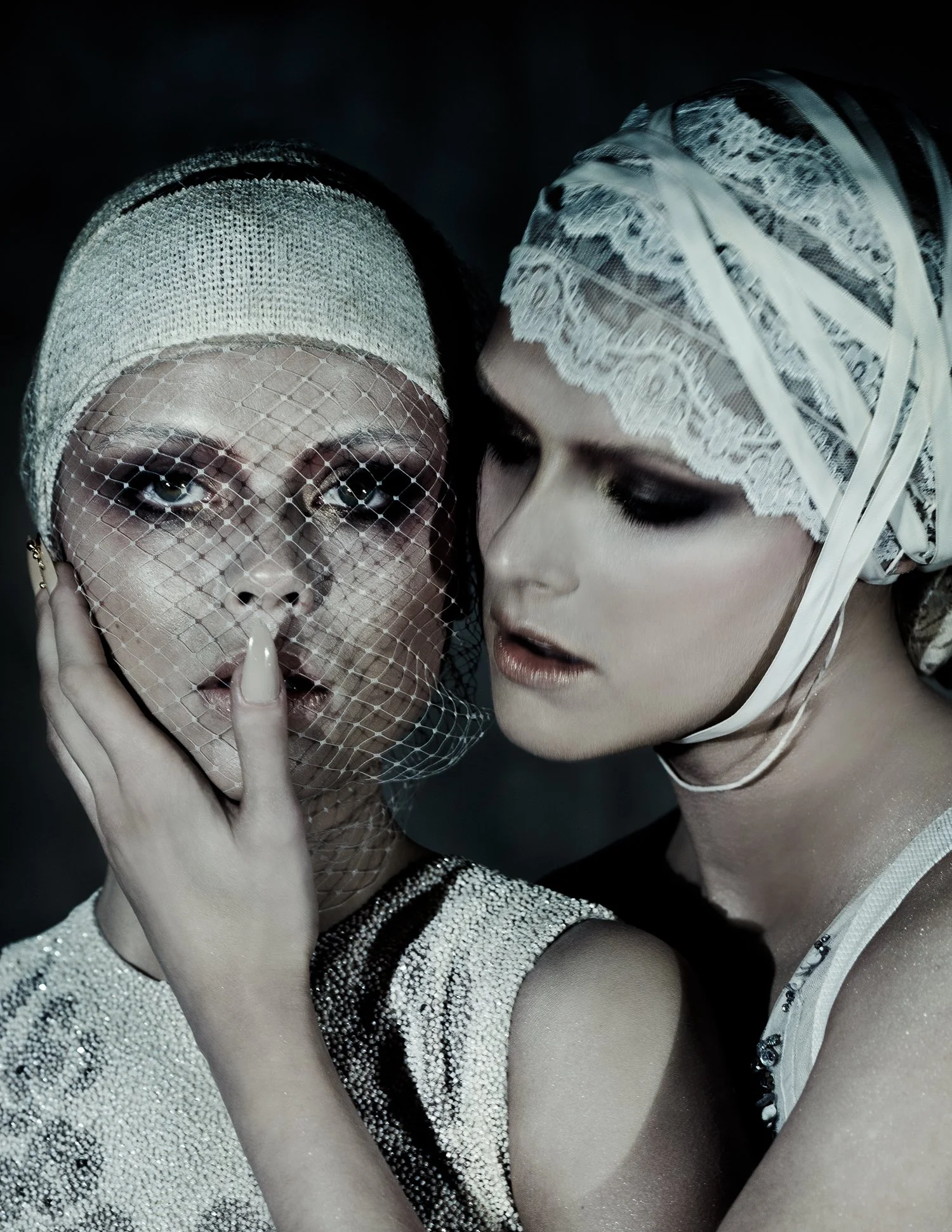 Two women with vintage headscarves and dark makeup, one touching her face, posing closely with serious expressions.