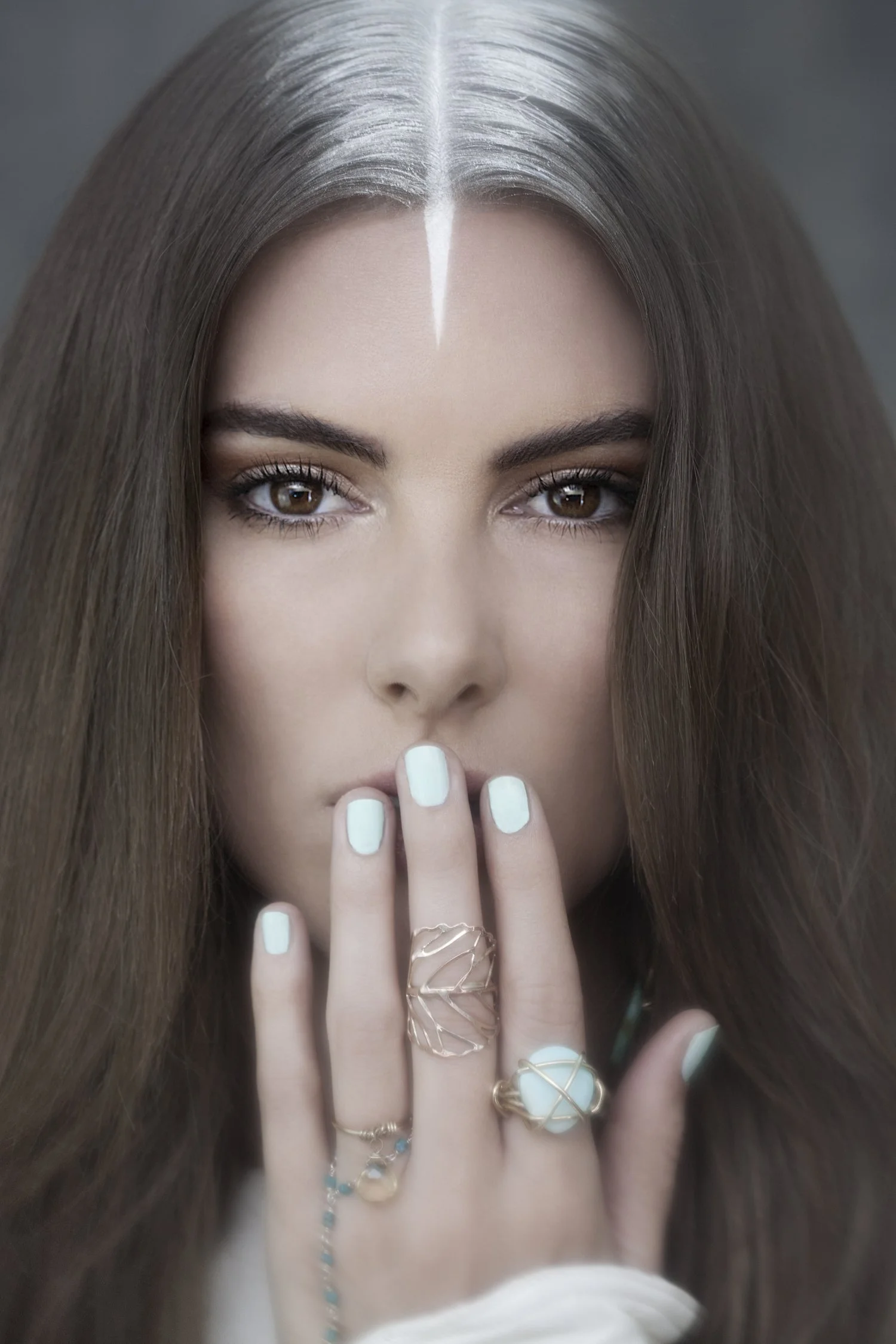 Close-up of a woman with long brown hair, brown eyes, and multiple rings on her fingers, covering her mouth with her hand.
