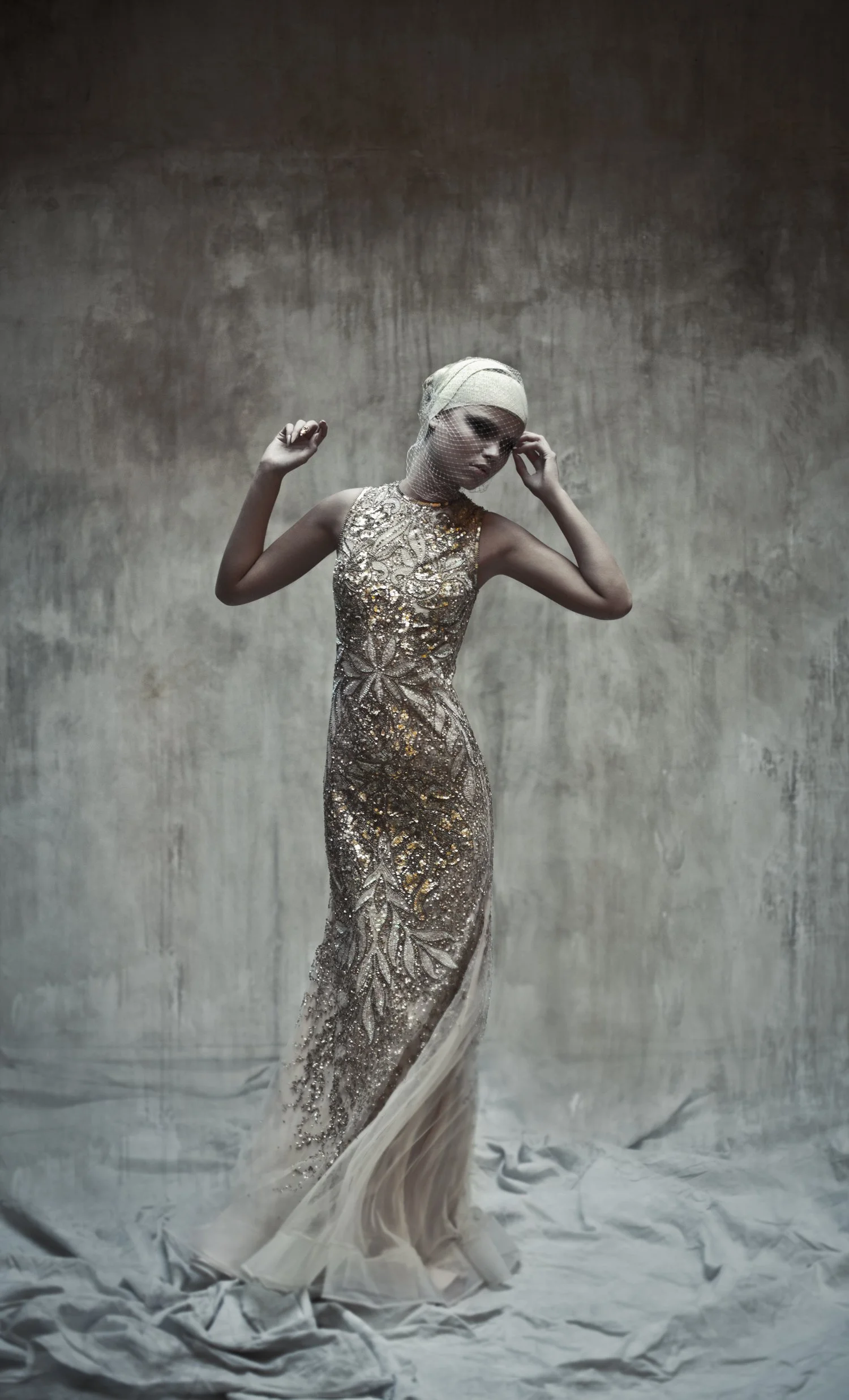 A woman wearing an elegant, gold and silver sequined evening gown with intricate floral patterns, standing against a textured gray background, with her head wrapped in a light-colored fabric and a netted veil over her face, striking a graceful pose.