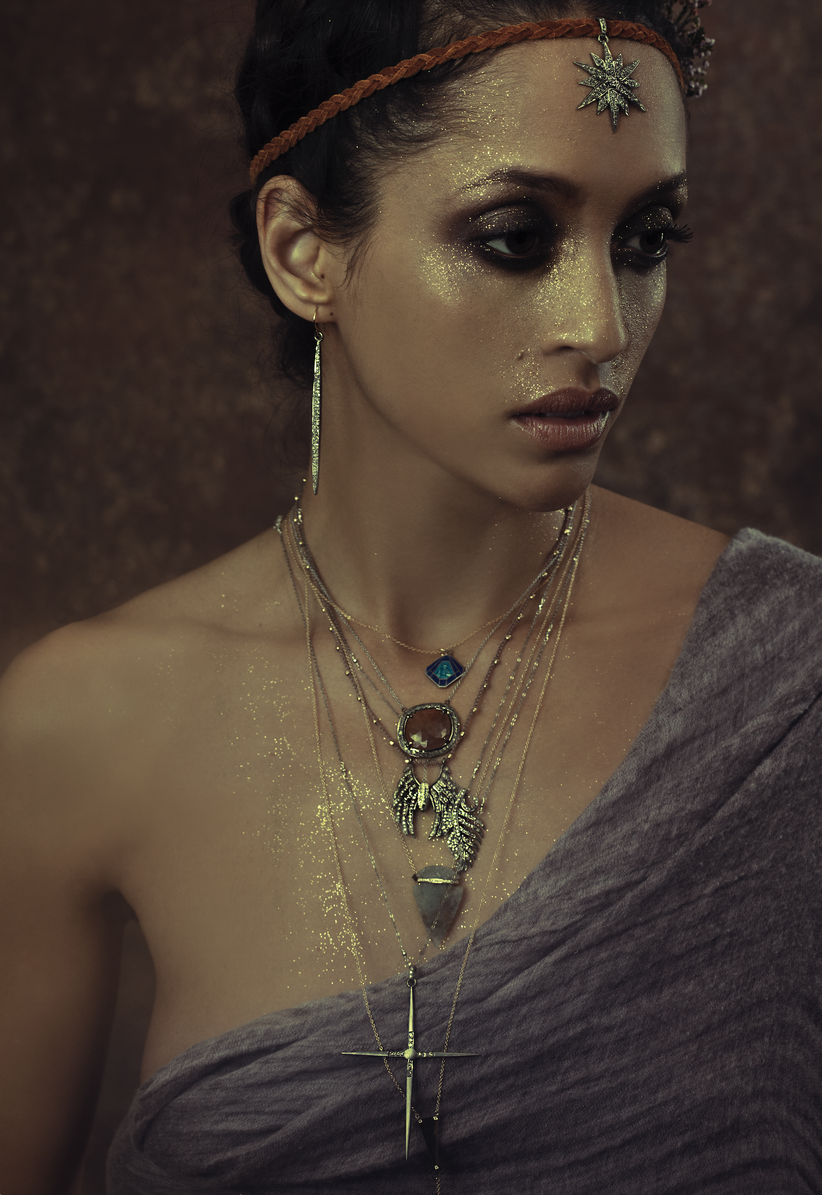A woman with dark hair and gold glitter on her face and shoulders, wearing multiple layered necklaces with various pendants, including a cross, feathers, and a gemstone, along with long earrings and a brown braided headband, dressed in a gray off-sho
