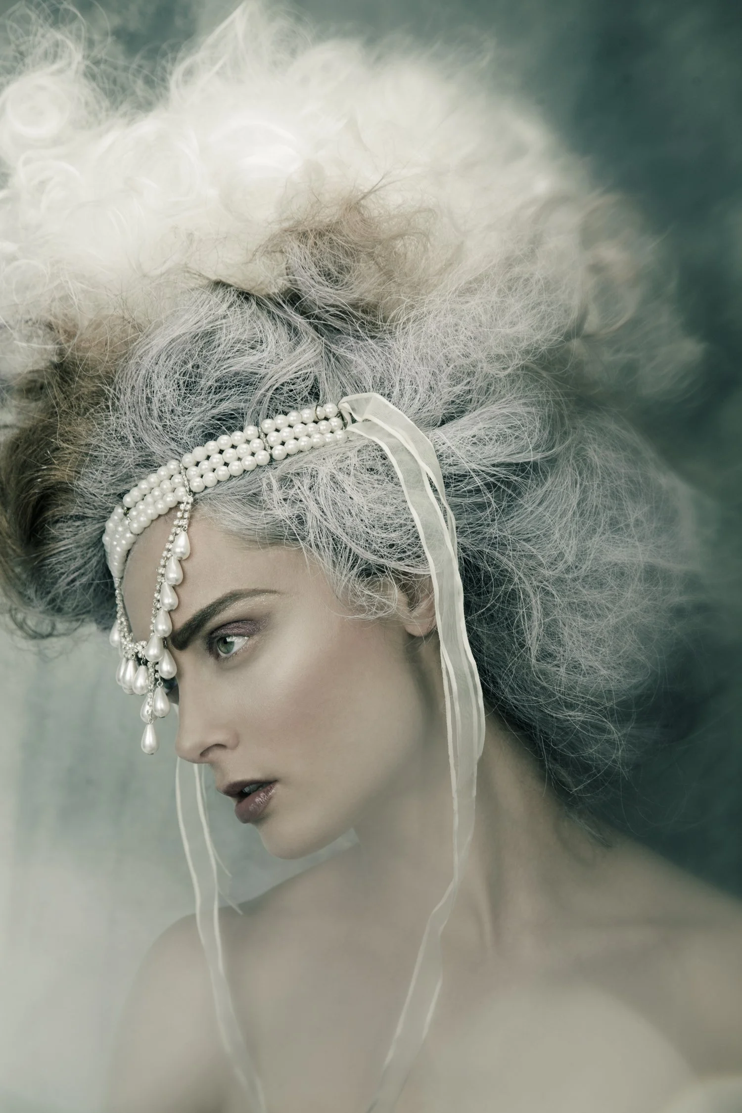 A person with elaborate, voluminous gray and white hair styled in an avant-garde fashion, wearing a pearl headband with dangling pearl embellishments, and have makeup with defined eyebrows and a serious expression.