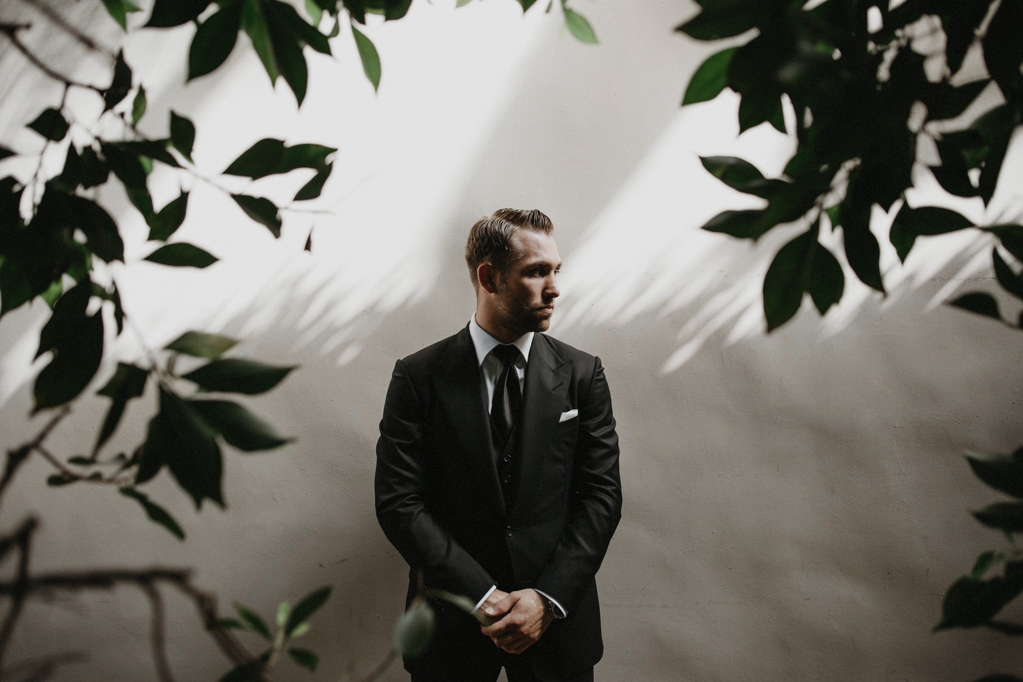 A handsome groom in a black suit stands against a white wall surrounded by trees at The Resort at Pelican Hill for wedding photos with j.olson weddings.