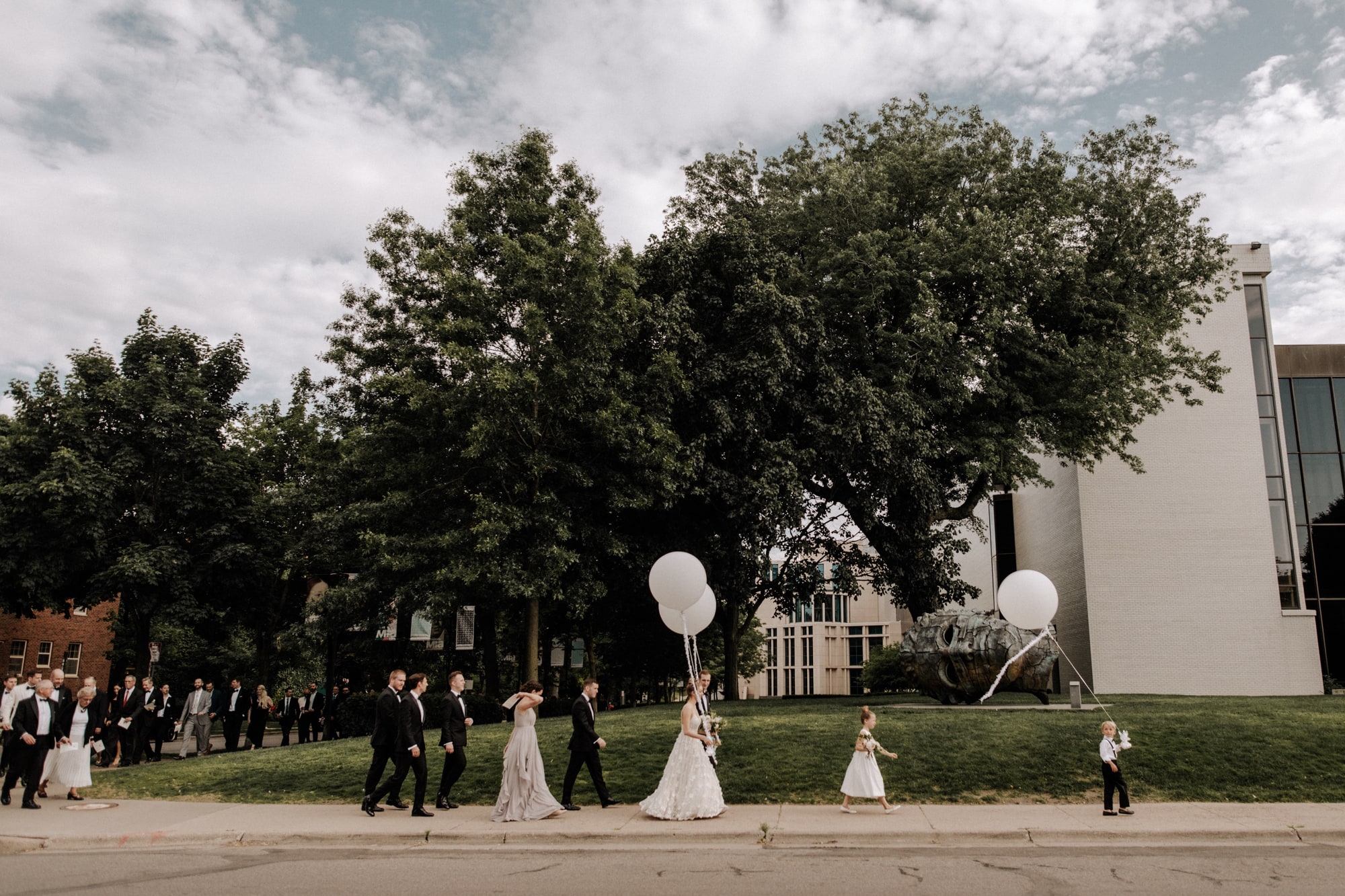 A bride and groom hold large white helium balloons while walking along the sidewalk with their wedding party outside of the Minneapolis Institute of Art.
