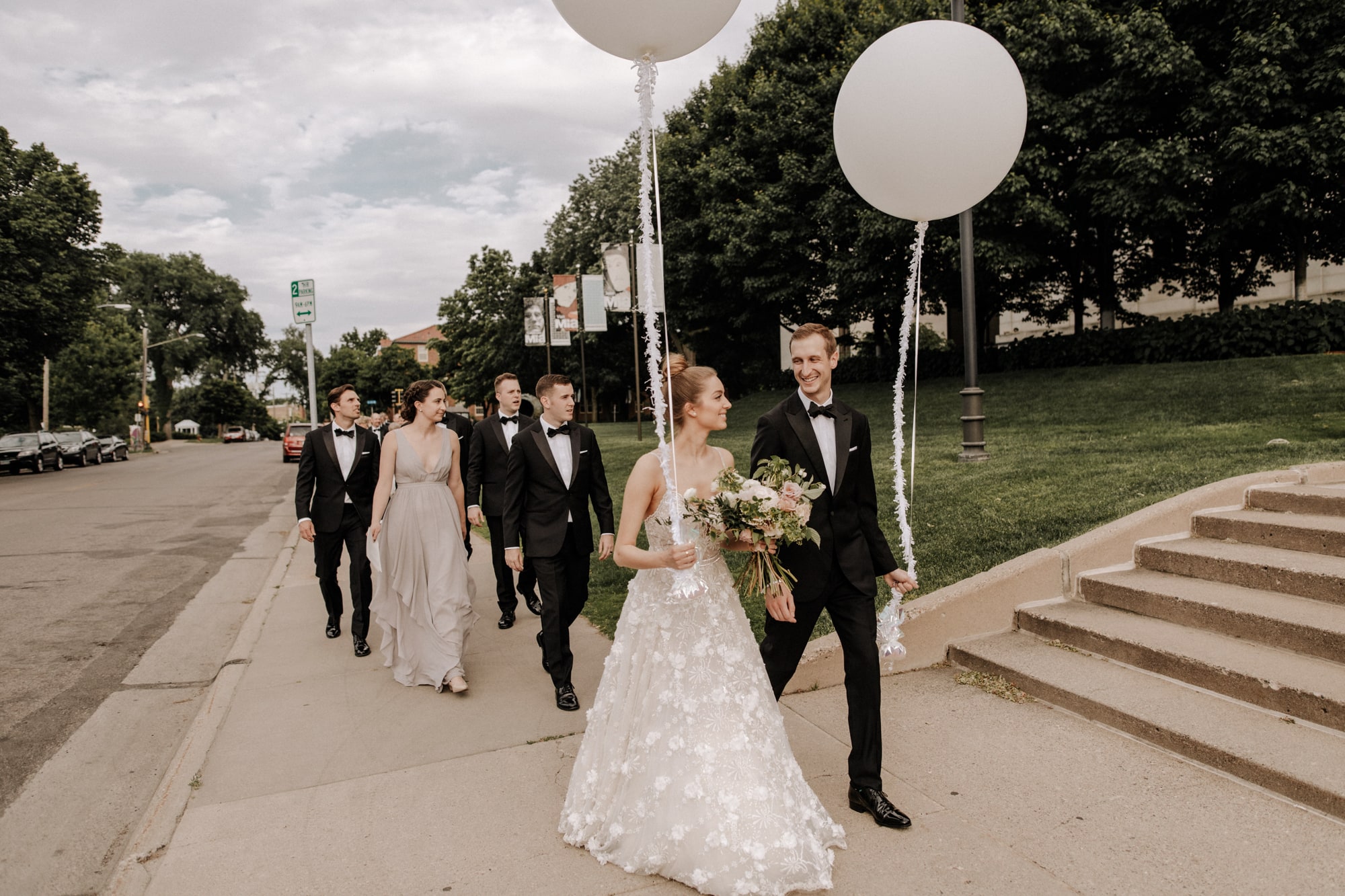 A newlywed bride and groom smile at each other while holding large white balloons and walking down the street with their wedding party in Minneapolis.