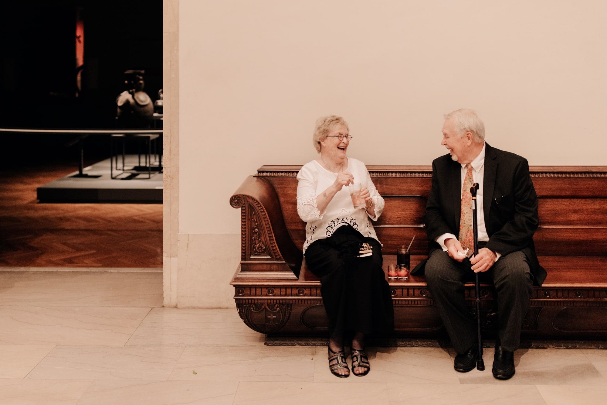 A senior couple laugh together while sitting on a wood bench during a wedding at the Minneapolis Institute of Art.