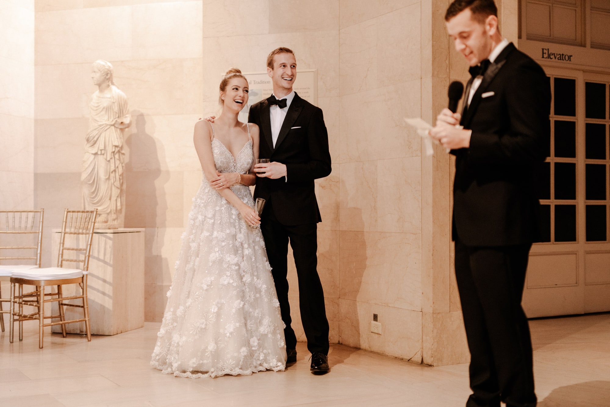 A bride and groom stand together, smiling, during speeches at their Minneapolis Institute of Art wedding. 