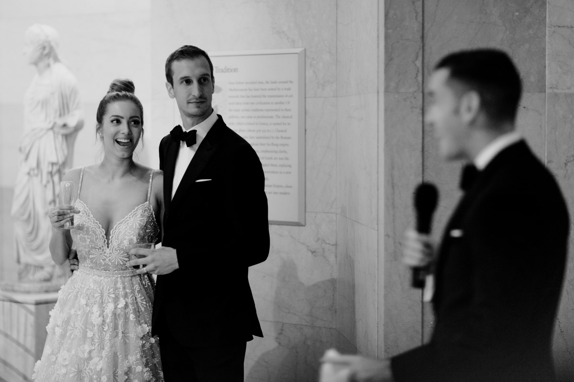 A beautiful bride and happy groom stand together, smiling and listening to wedding speeches inside the Minneapolis Institute of Art.