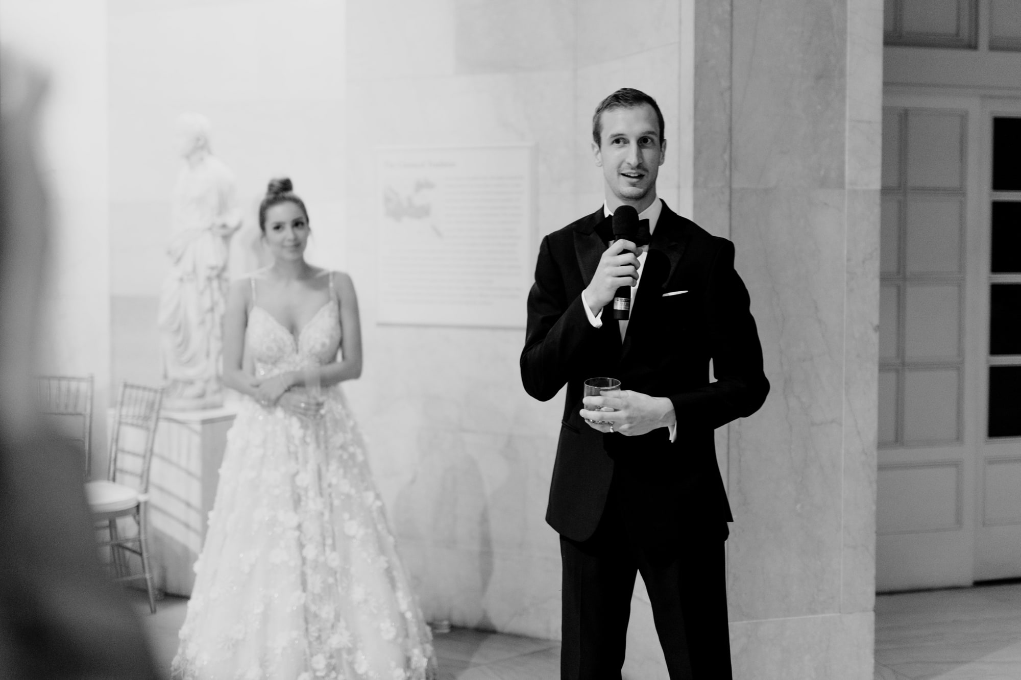 A groom speaks into a microphone, his bride standing behind him, during their Minneapolis Institute of Art wedding.