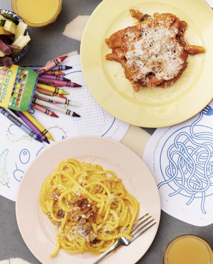 Prato de massa com molho de tomate e queijo, presunto na folha de papel de desenho, brinquedos de crayola e copos de suco ao redor.