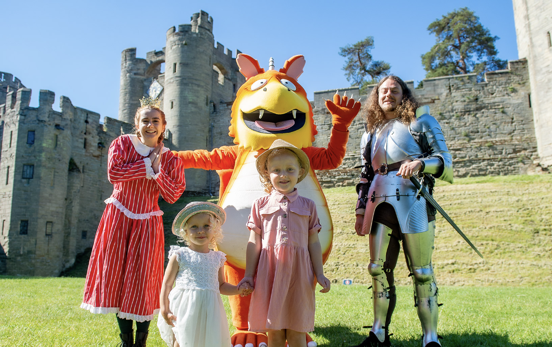 Família com crianças e adultos em fantasia de personagens de contos de fada, na frente de um castelo de pedra em dia ensolarado.