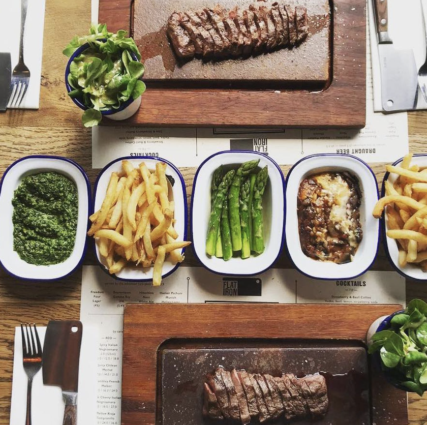 Pratos preparados com picanha, molho verde, batatas fritas, aspargos e molho de berinjela gratinado numa mesa de restaurante.