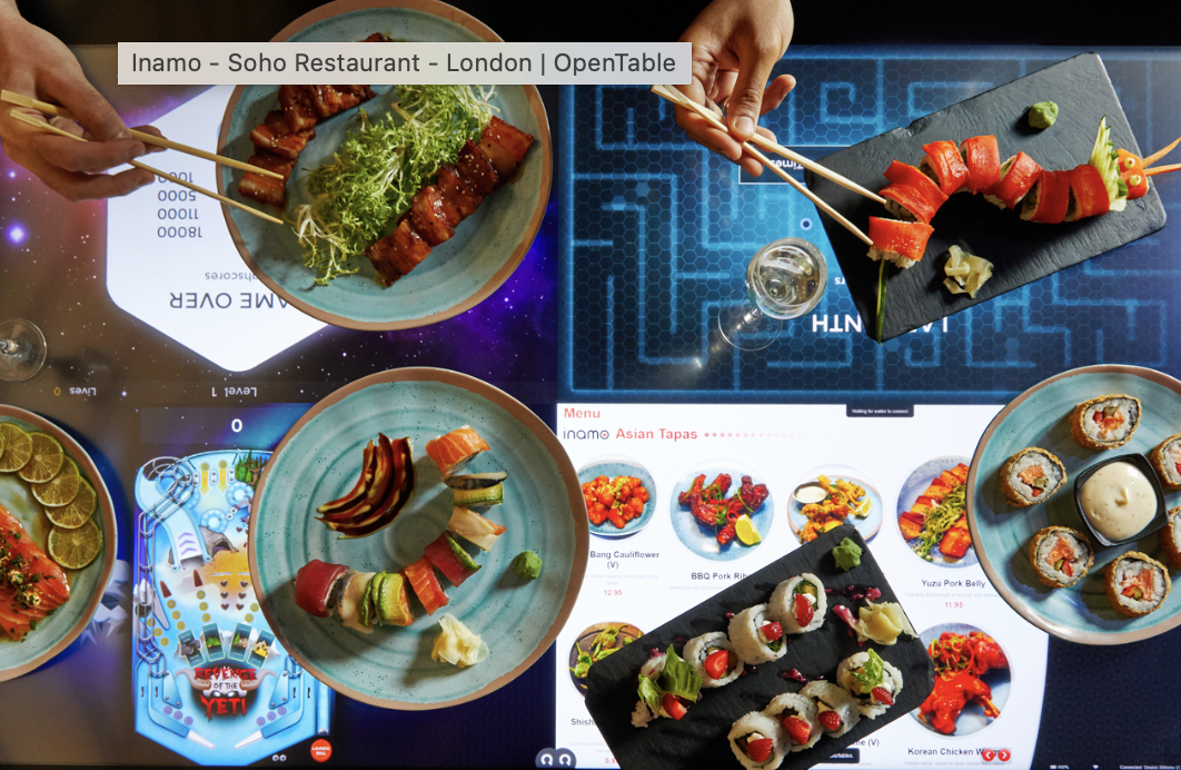 Variedade de pratos de comida japonesa, incluindo sushis, sashimis, rolinhos e espetinhos, dispostos sobre mesas com telas digitais exibindo menus de restaurante.