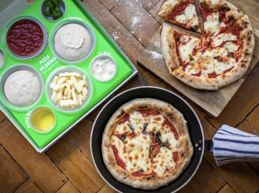 Duas pizzas de queijo em uma bancada de madeira, uma em uma frigideira e outra em uma tábua de madeira, acompanhadas de ingredientes para pizza e temperos em uma bandeja verde ao lado.
