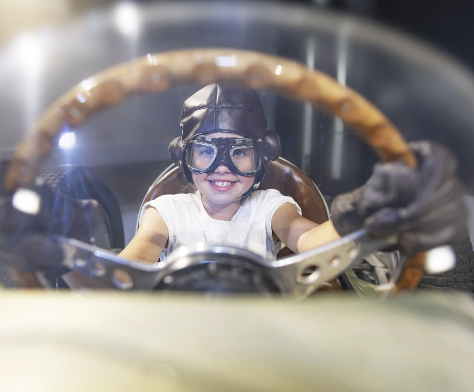 Menina sorridente usando capacete, óculos de proteção e roupas de piloto de corrida, sentado em um carro de corrida vintage.