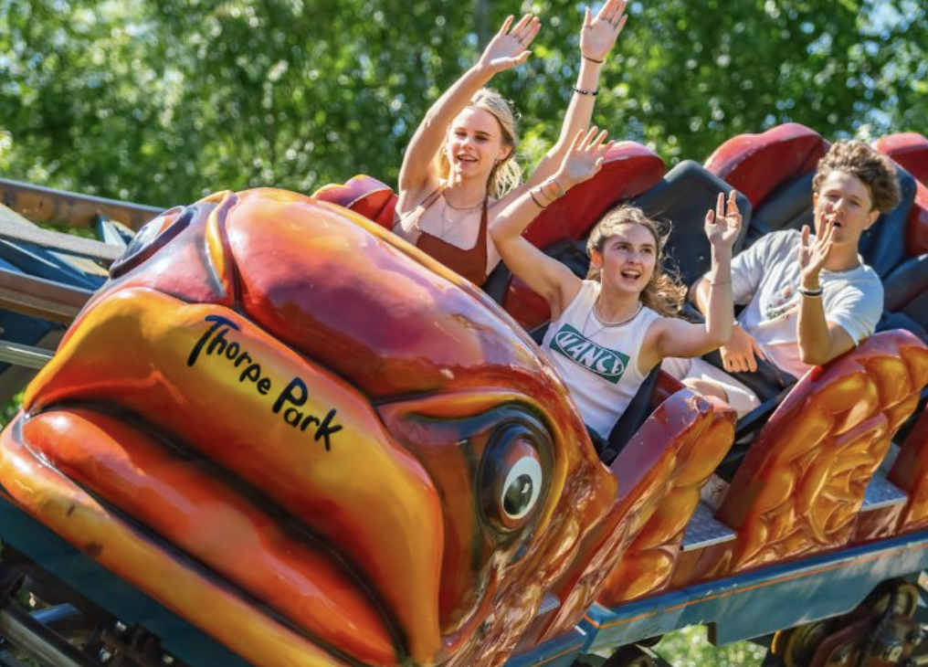 Três pessoas se divertindo em uma montanha-russa com um tema de caranguejo, na entrada com o nome Thorpe Park, rodeadas por árvores verdes.
