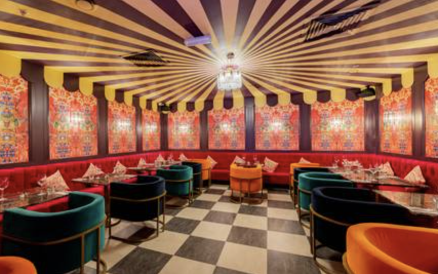 Interior de restaurante luxuoso com teto decorado com linhas inclinadas, luz central, paredes com papel de parede floral, cadeiras verdes e laranjas, paredes vermelhas e piso de xadrez preto e branco.