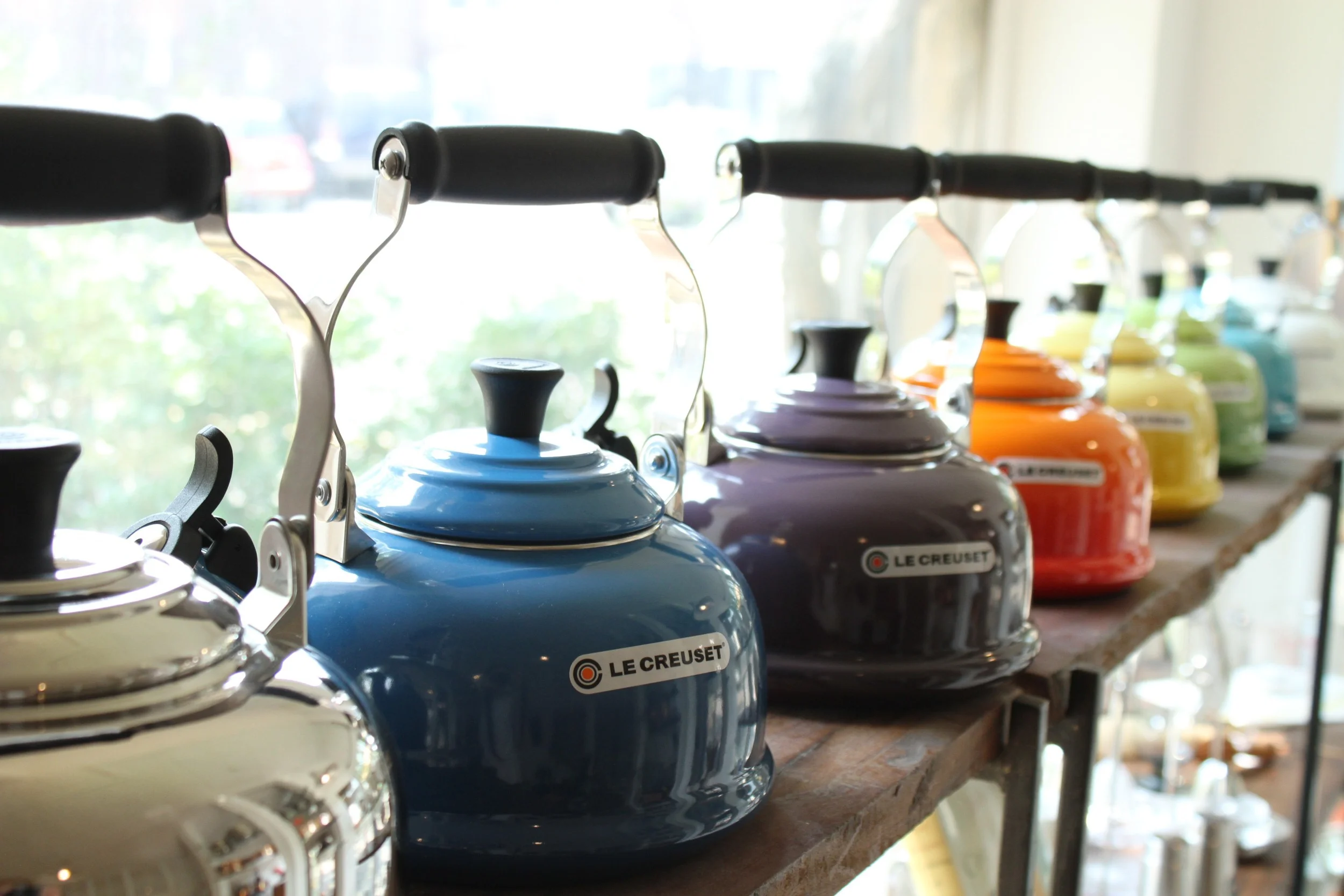 Le Creuset 1.8 qt whistling tea kettles , pictured from front to back in stainless steel, Marseille, Cassis, Flame, Soleil, Palm, Carribean, and White. Also available in Cherry.