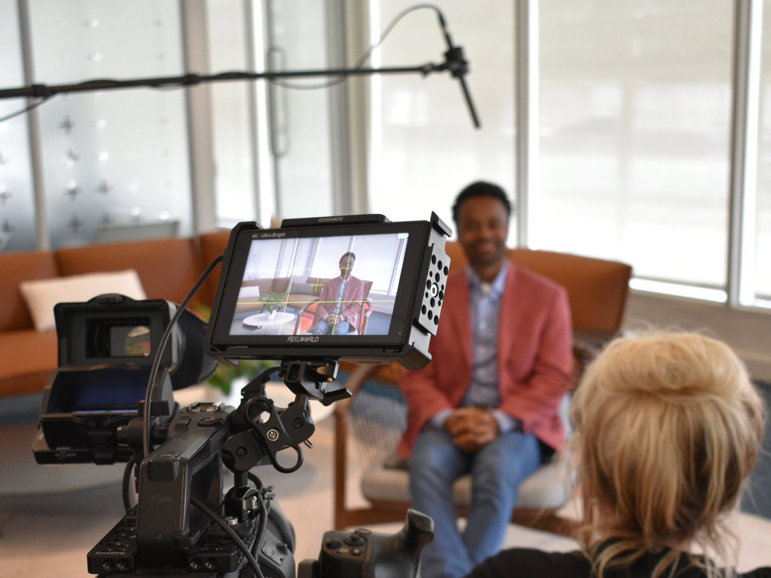An on camera interview: a monitor frames a smiling man in a red blazer while a producer watches