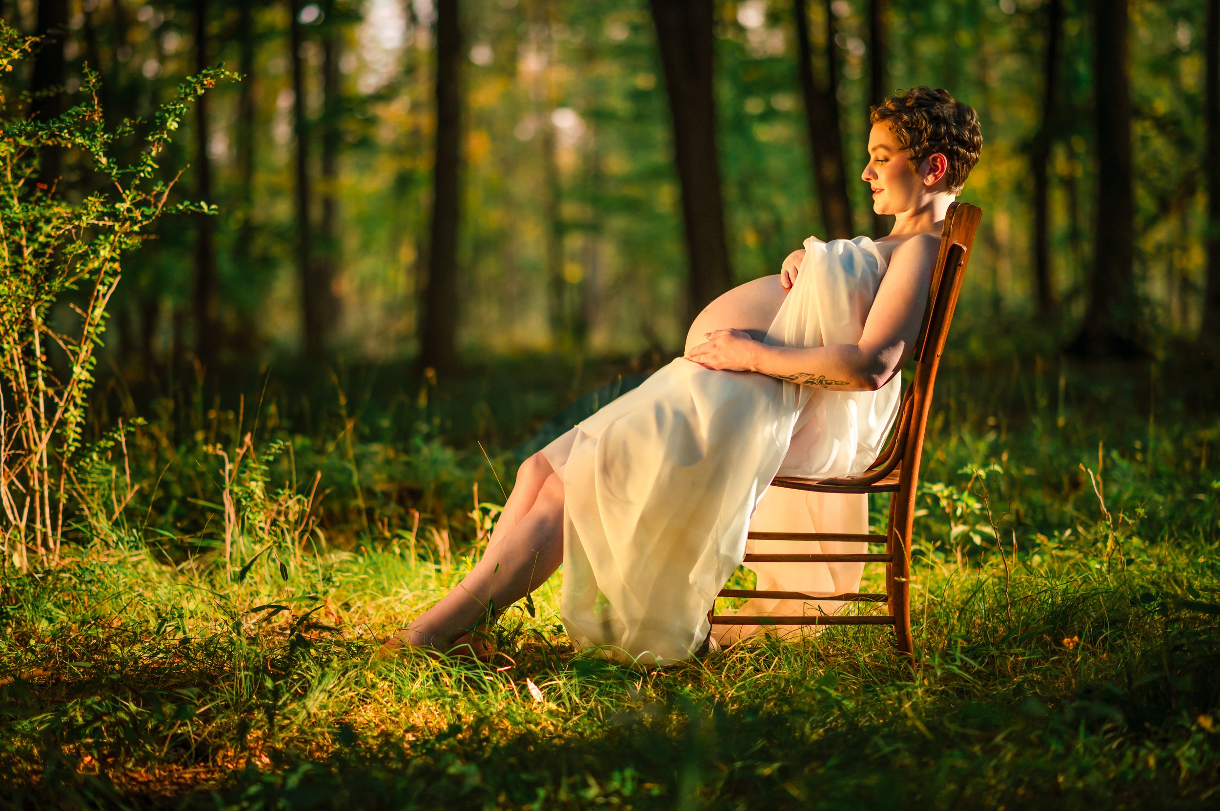 A woman in a white dress sitting on a wooden chair in a forest, illuminated by warm sunlight.