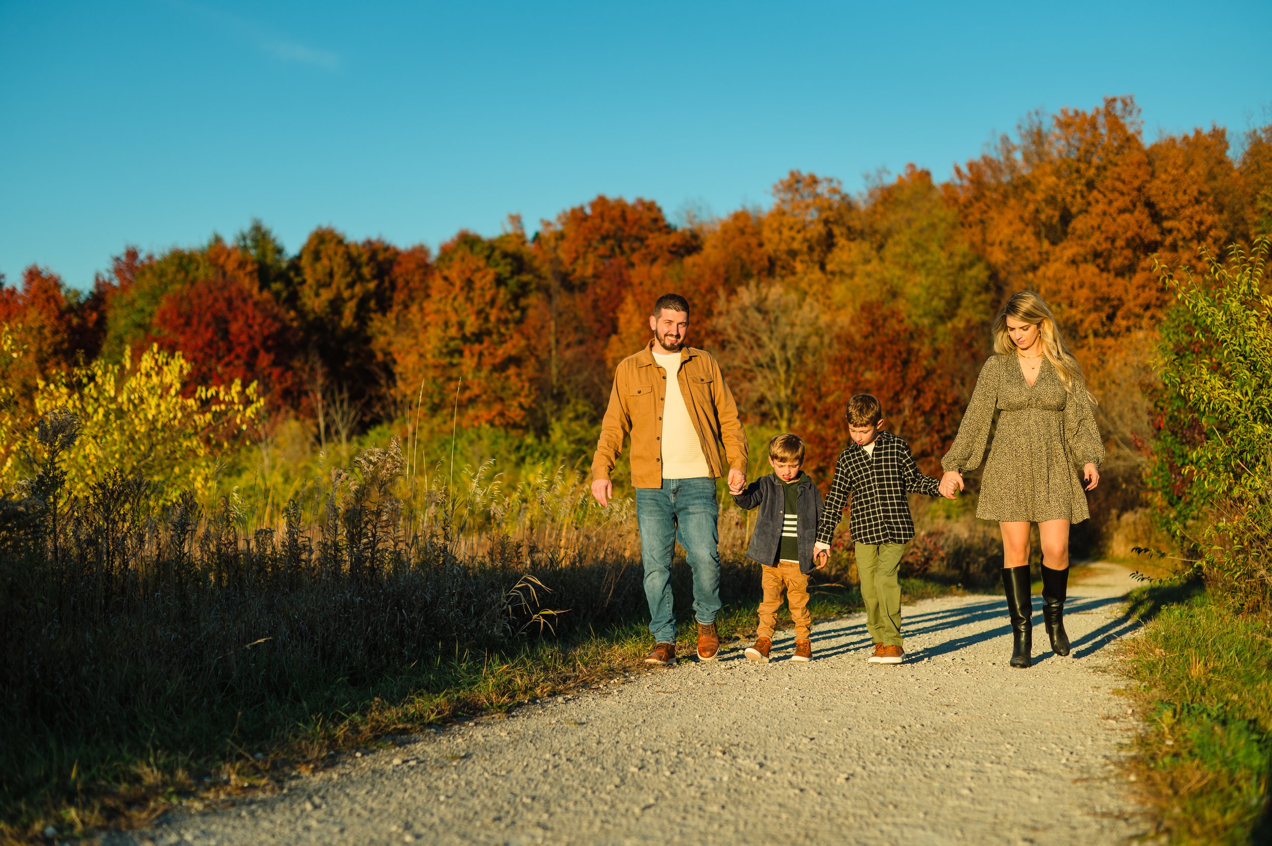 Family of four walking on a gravel path during autumn, with colorful trees and a clear blue sky in the background.