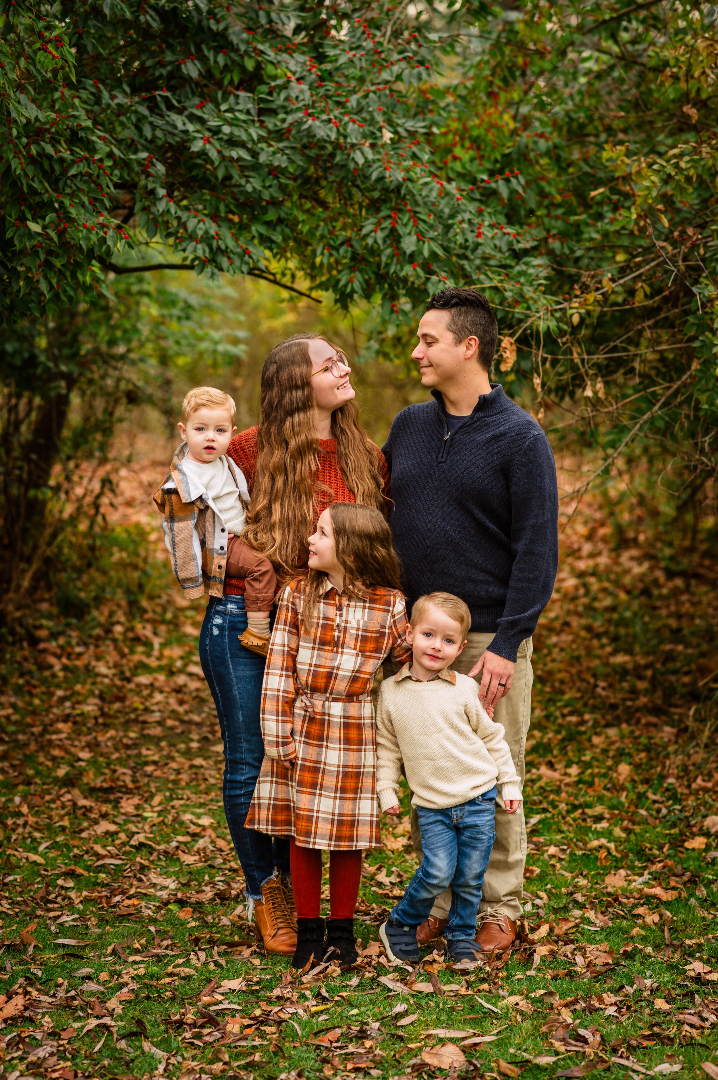 A happy family of five, including two young children, stands outdoors in a wooded area with fall leaves on the ground and green foliage overhead.