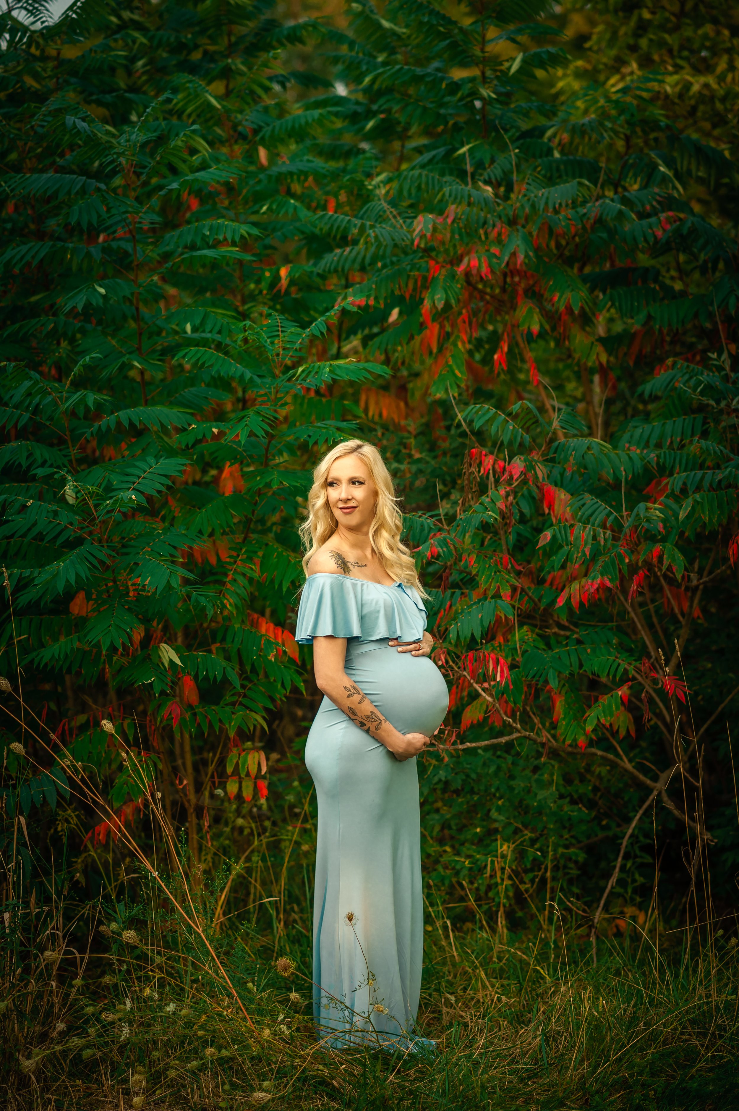 Pregnant woman in a light blue dress standing in front of green and red foliage, holding her belly with both hands.