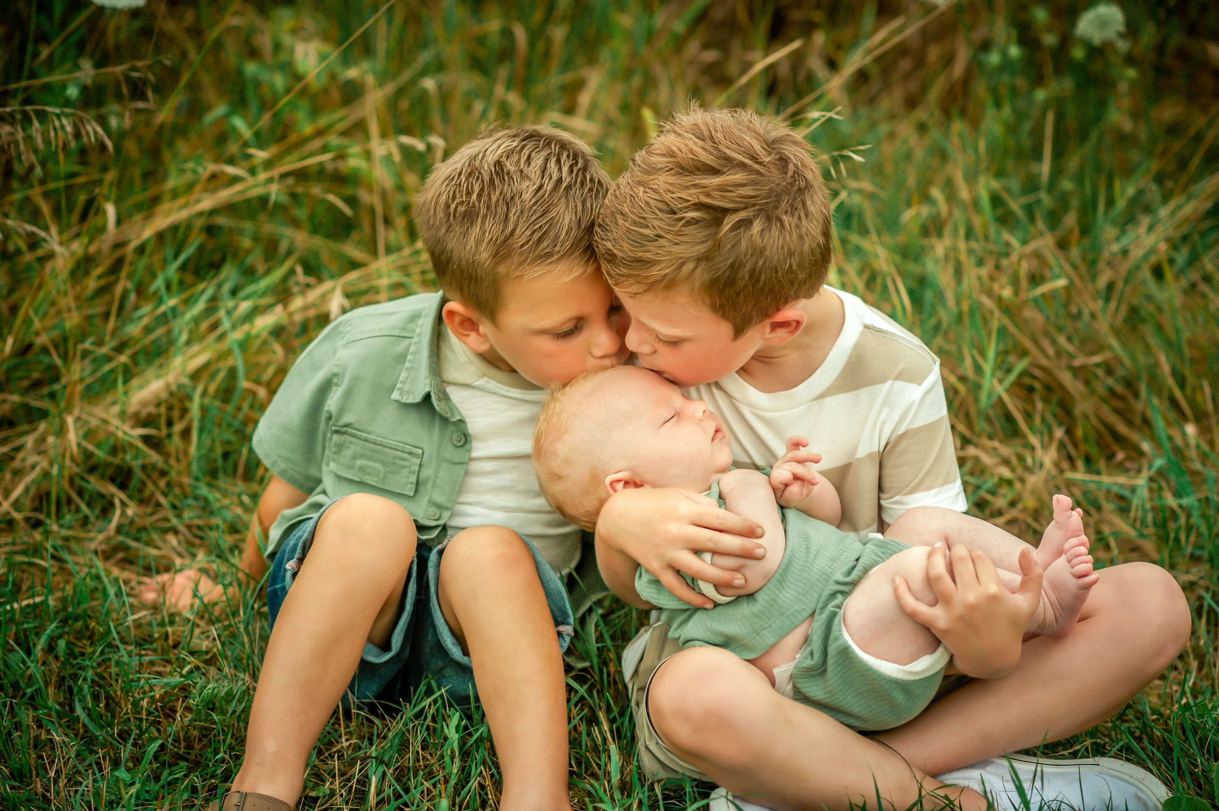 Three young boys, two older and one baby, sitting outdoors on grass, kissing and holding the baby