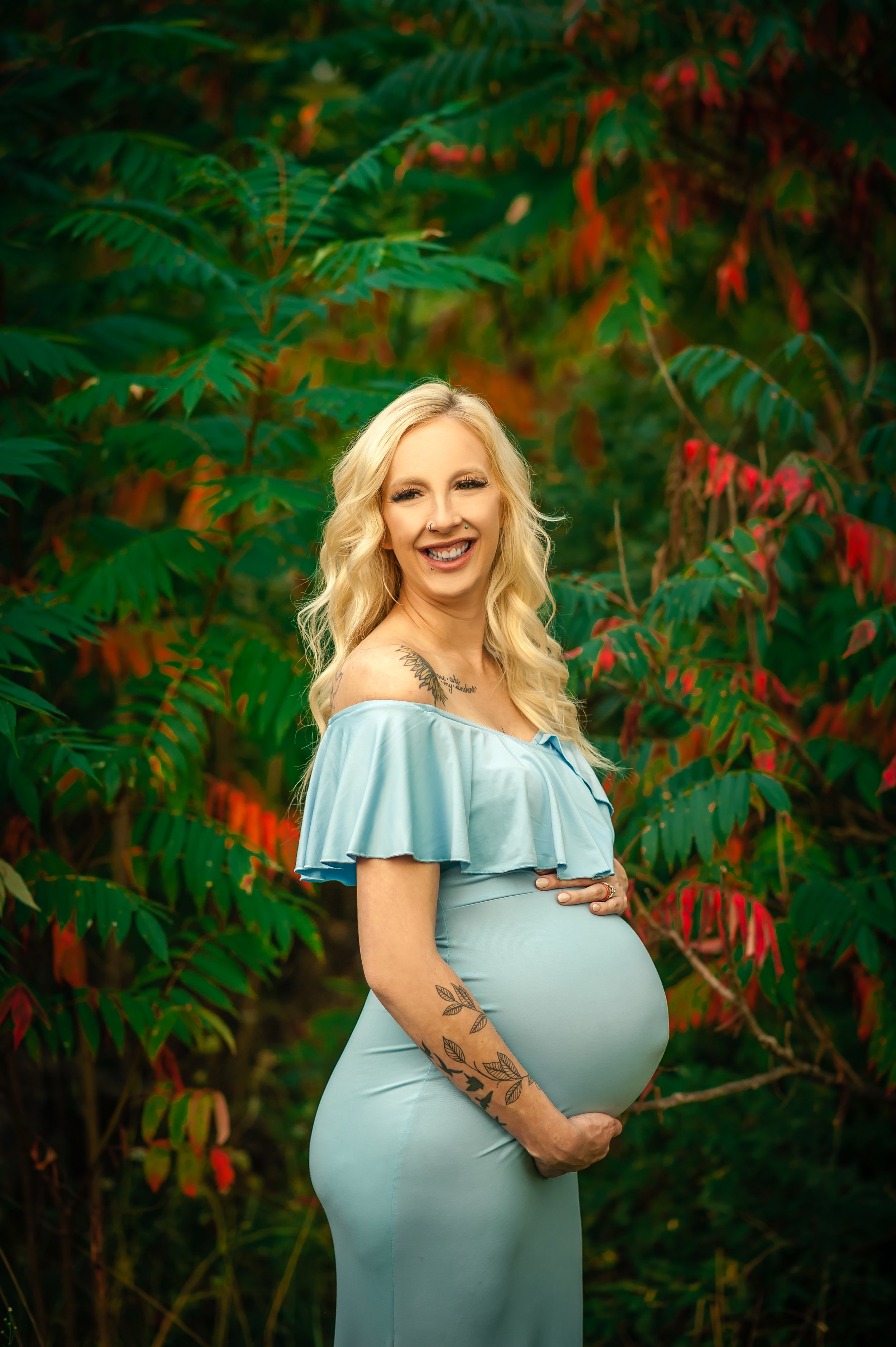 A pregnant woman with long blonde hair, tattoos on her arm and shoulder, smiling and holding her belly, standing outdoors in front of green and red foliage, wearing a light blue off-shoulder dress.