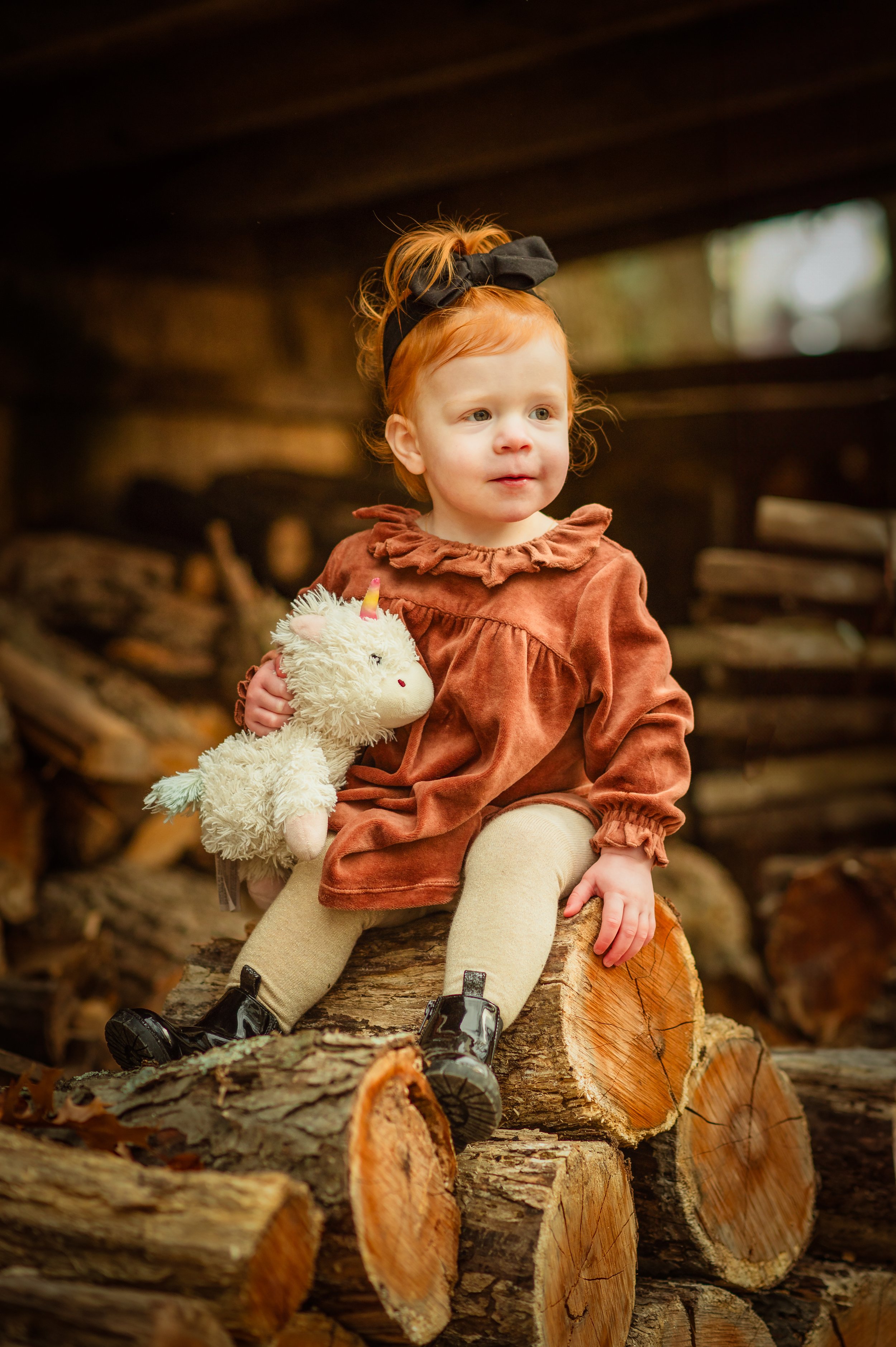 A young girl with red hair and a black bow, wearing a brown velvet dress, beige tights, and black shoes, sitting on a pile of logs indoors, holding a plush unicorn toy.