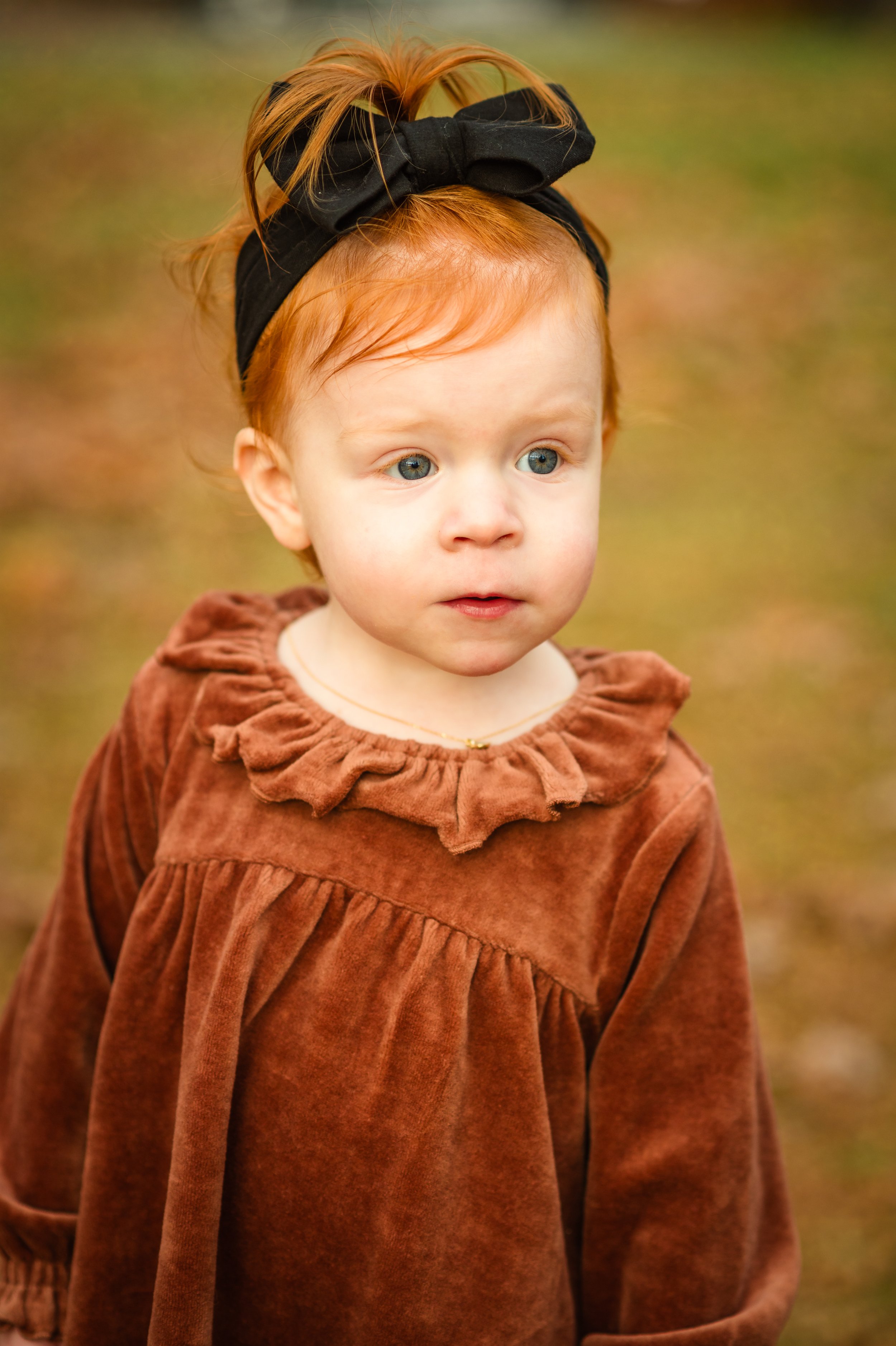 A young girl with red hair and blue eyes standing outdoors with a blurred foliage background, wearing a brown dress with ruffles and a black headband with a bow.