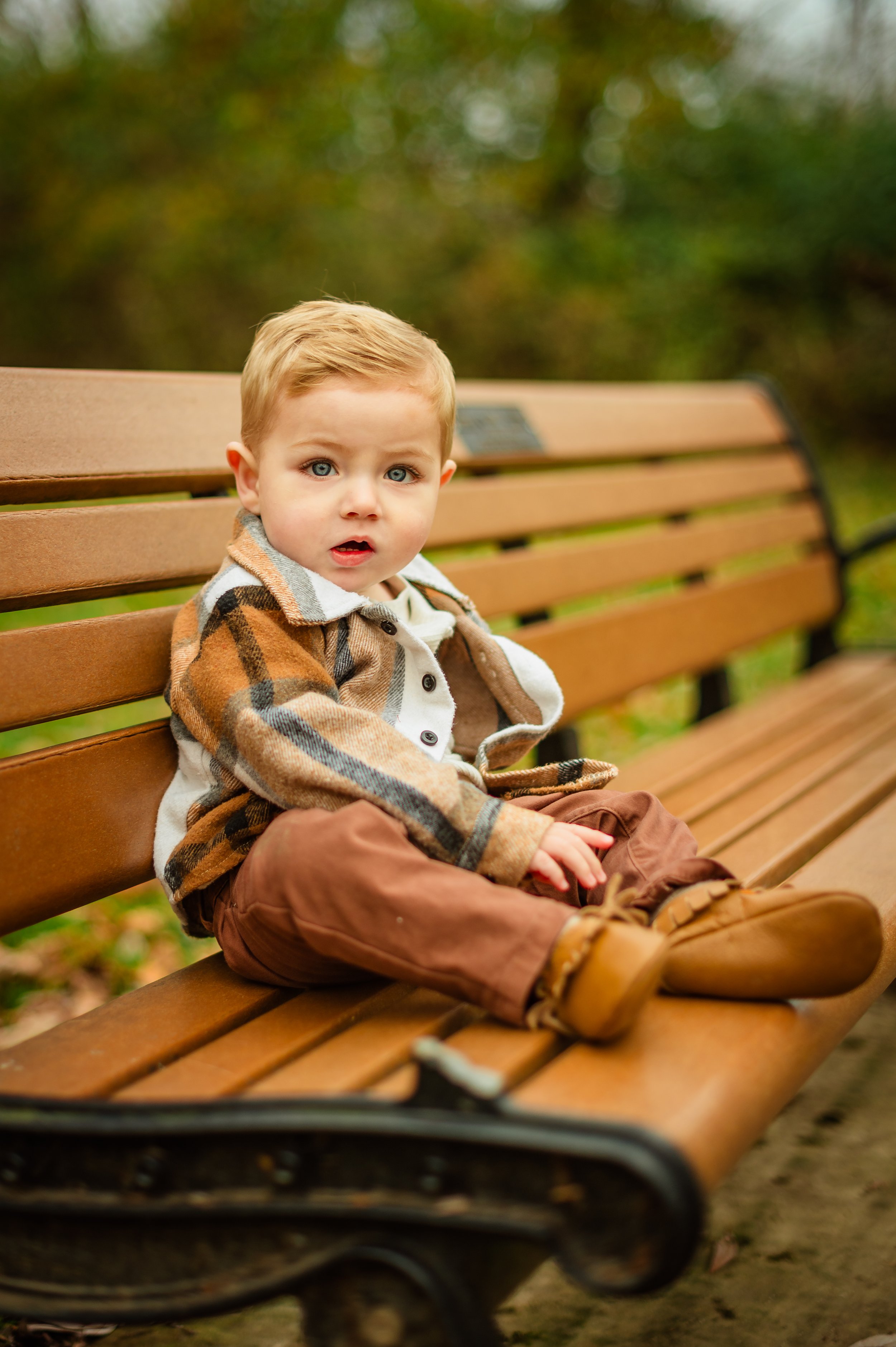 A young boy with blonde hair and blue eyes sitting on a wooden park bench in autumn, wearing a plaid jacket and brown pants.