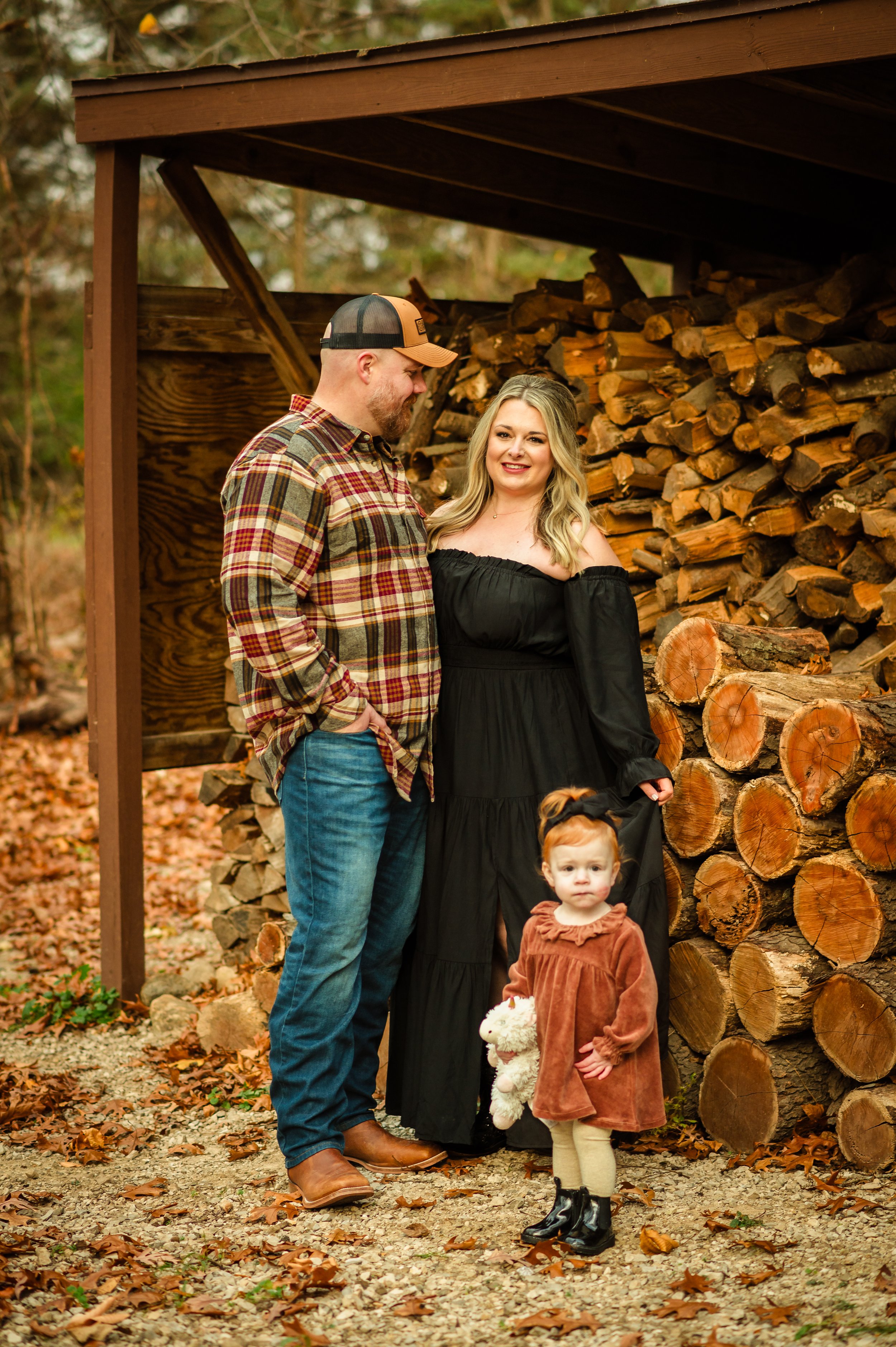 A family of three stands outdoors near a large pile of stacked firewood, with fallen leaves on the ground. The man is wearing a plaid shirt and jeans, the woman is in a black off-shoulder dress, and the young girl is dressed in a brown coat and holdi