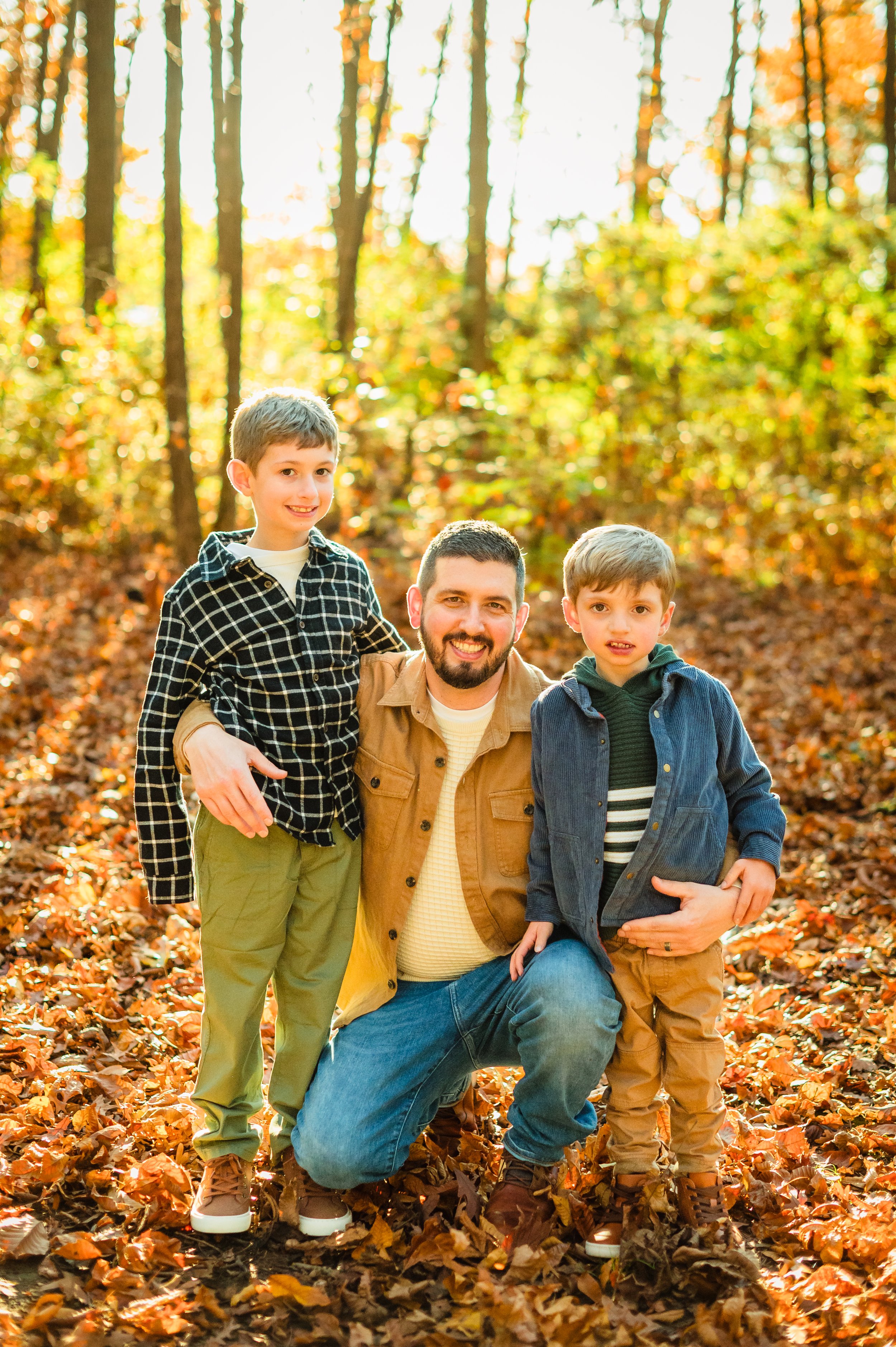 A man and two young boys in an autumn forest, surrounded by orange and brown fallen leaves, smiling for a photo.