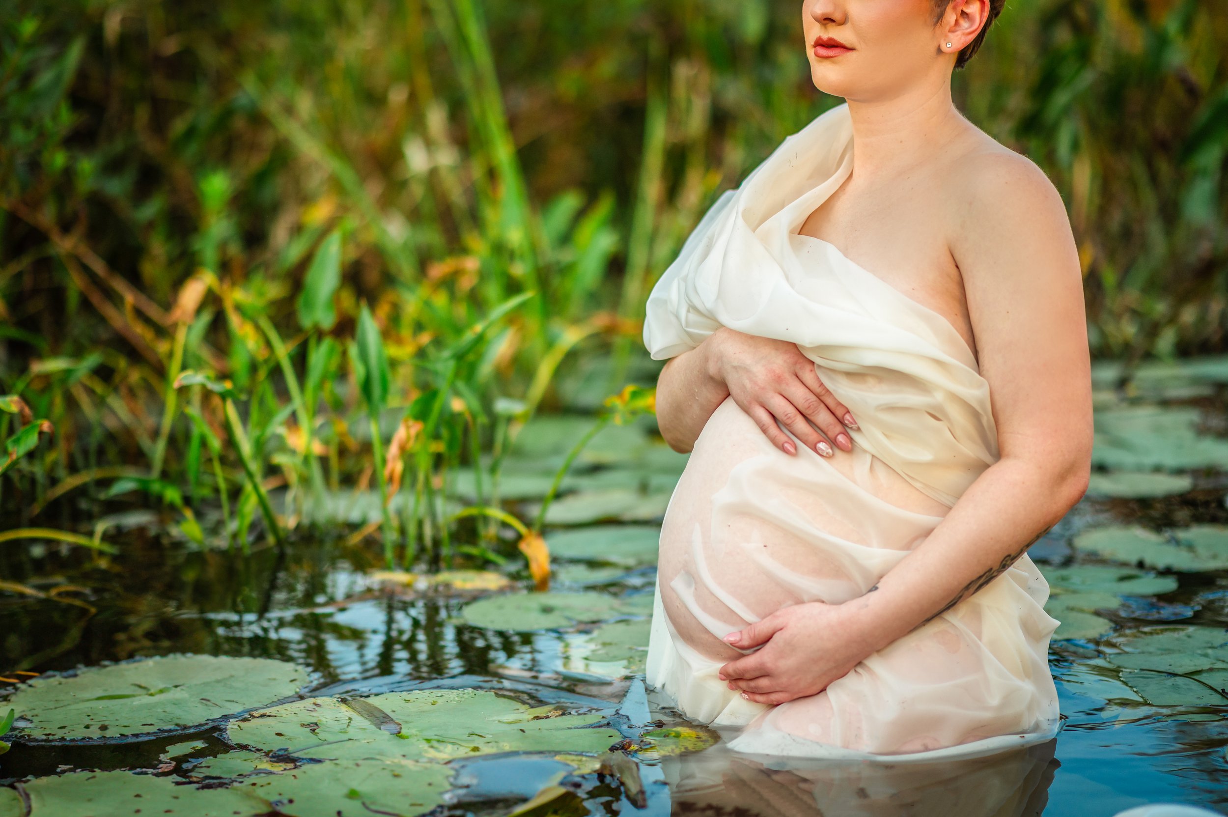 A pregnant woman standing in a pond surrounded by lilies and green plants, wearing a cream-colored fabric draped around her.
