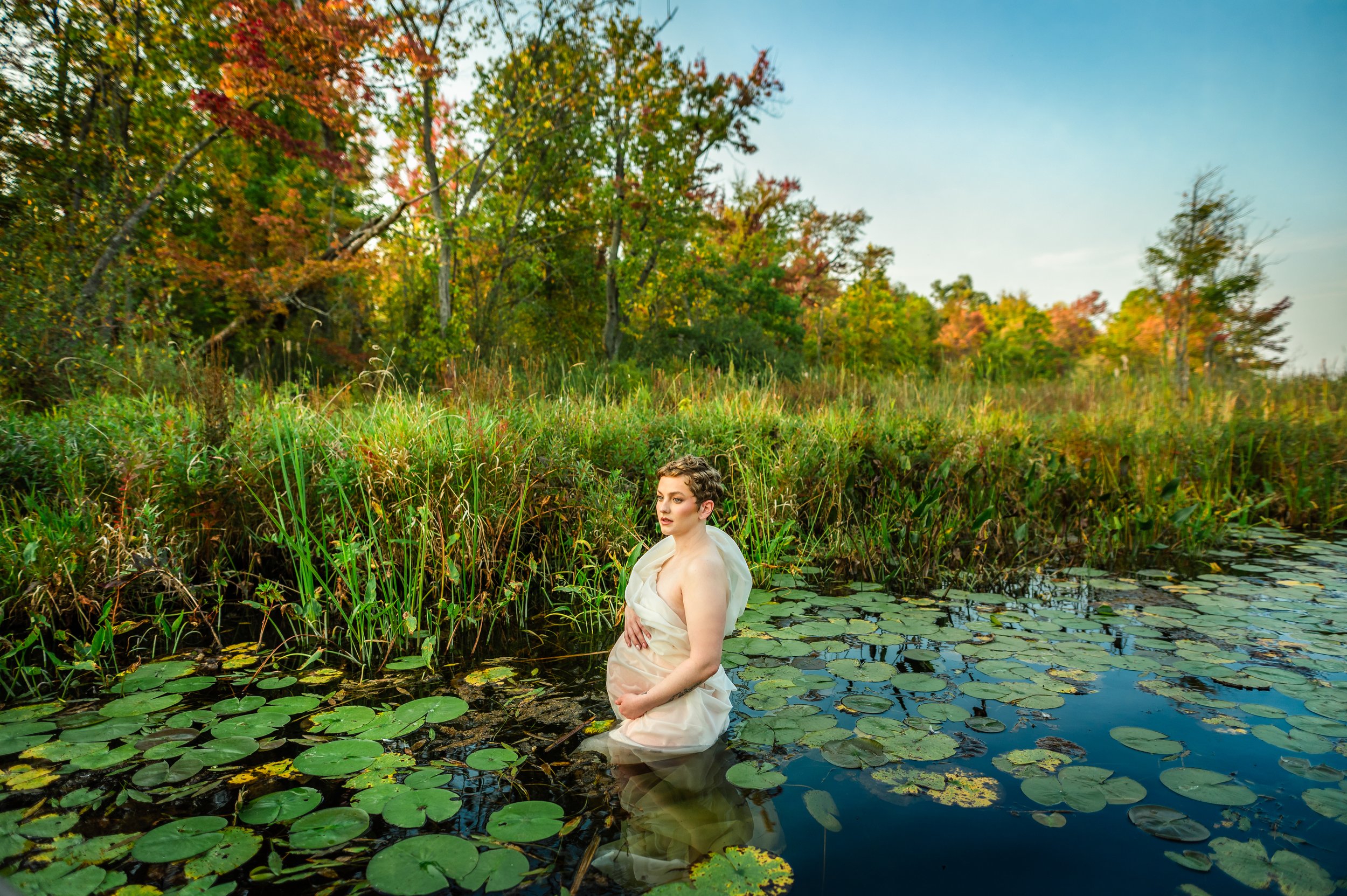 A pregnant woman in a flowing white dress kneels in a pond surrounded by lily pads and tall grass, with colorful autumn trees in the background.