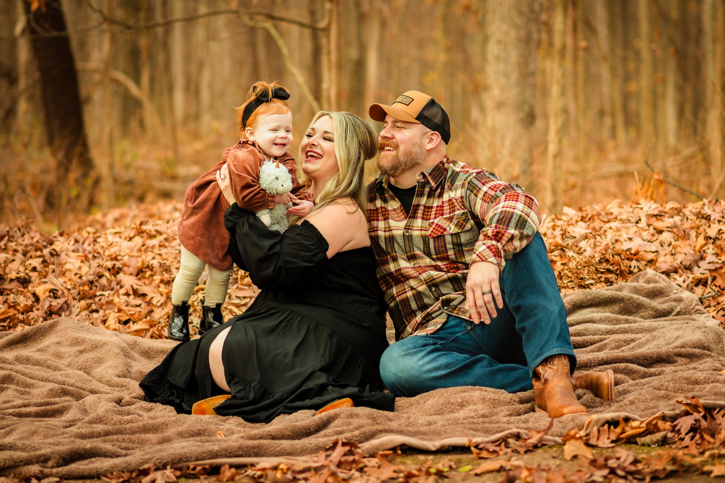 A family of three, a mother, father, and young daughter, enjoying a moment together in a fall forest, sitting on a blanket amid fallen leaves. The mother and daughter are smiling and laughing, and the father is watching happily.