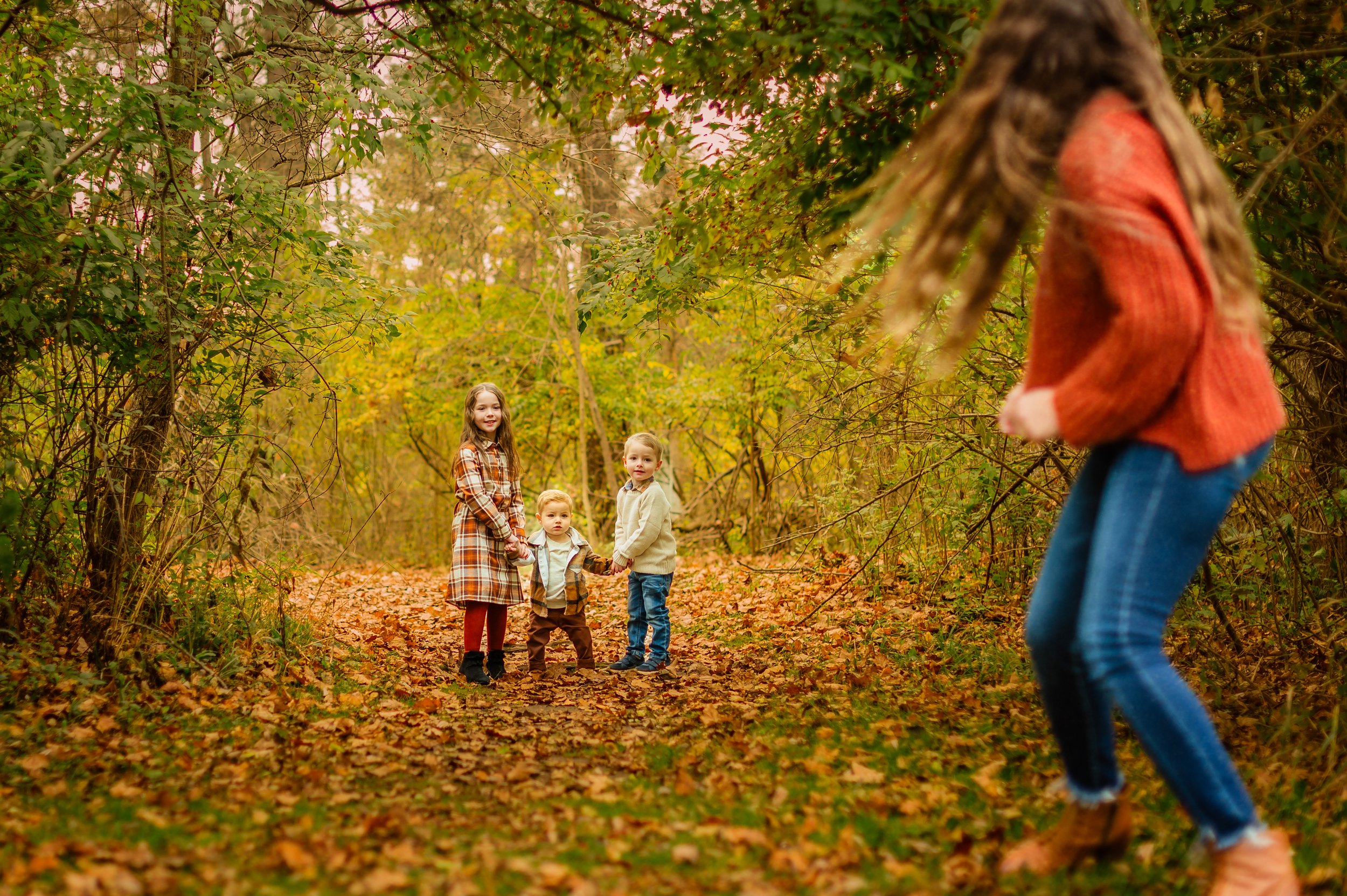 A woman with long hair in an orange sweater and jeans appears to be falling or tripping in a forest with fallen leaves. In the background, three children, a girl and two boys, are holding hands and smiling, dressed in autumn clothing, standing on a l
