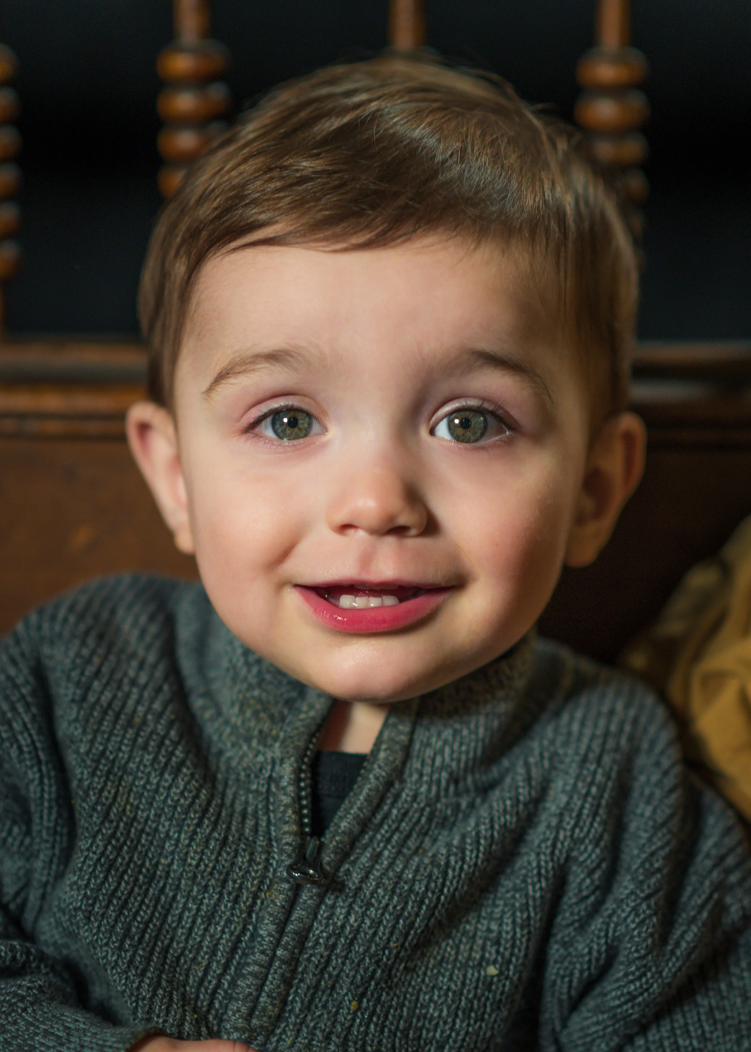 Close-up of a young boy with short brown hair and green eyes, smiling and wearing a gray zip-up sweater.