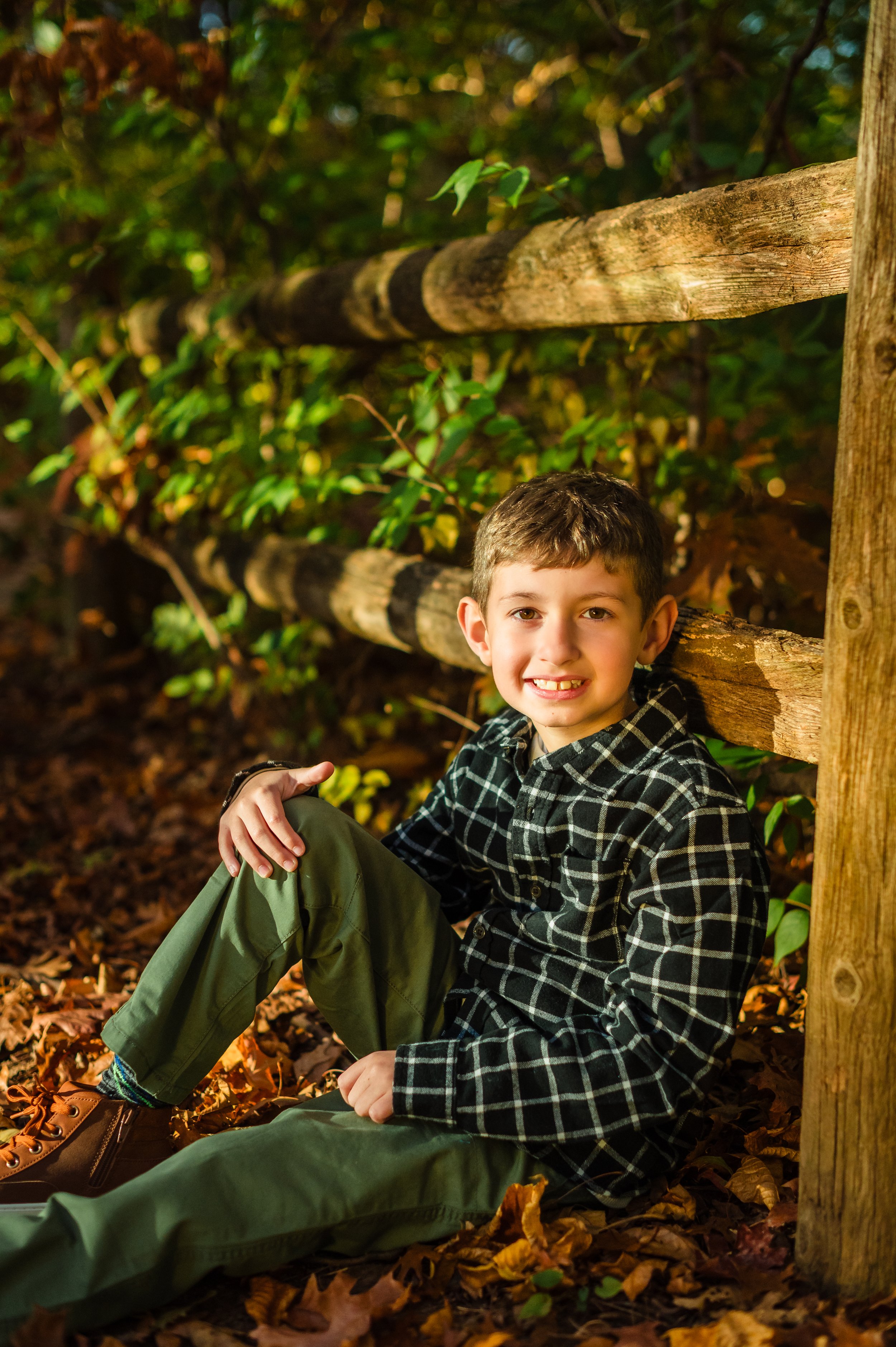 A young boy with short brown hair, wearing a black plaid shirt and green pants, sitting on the ground covered with autumn leaves, leaning against a wooden fence in a forested area.