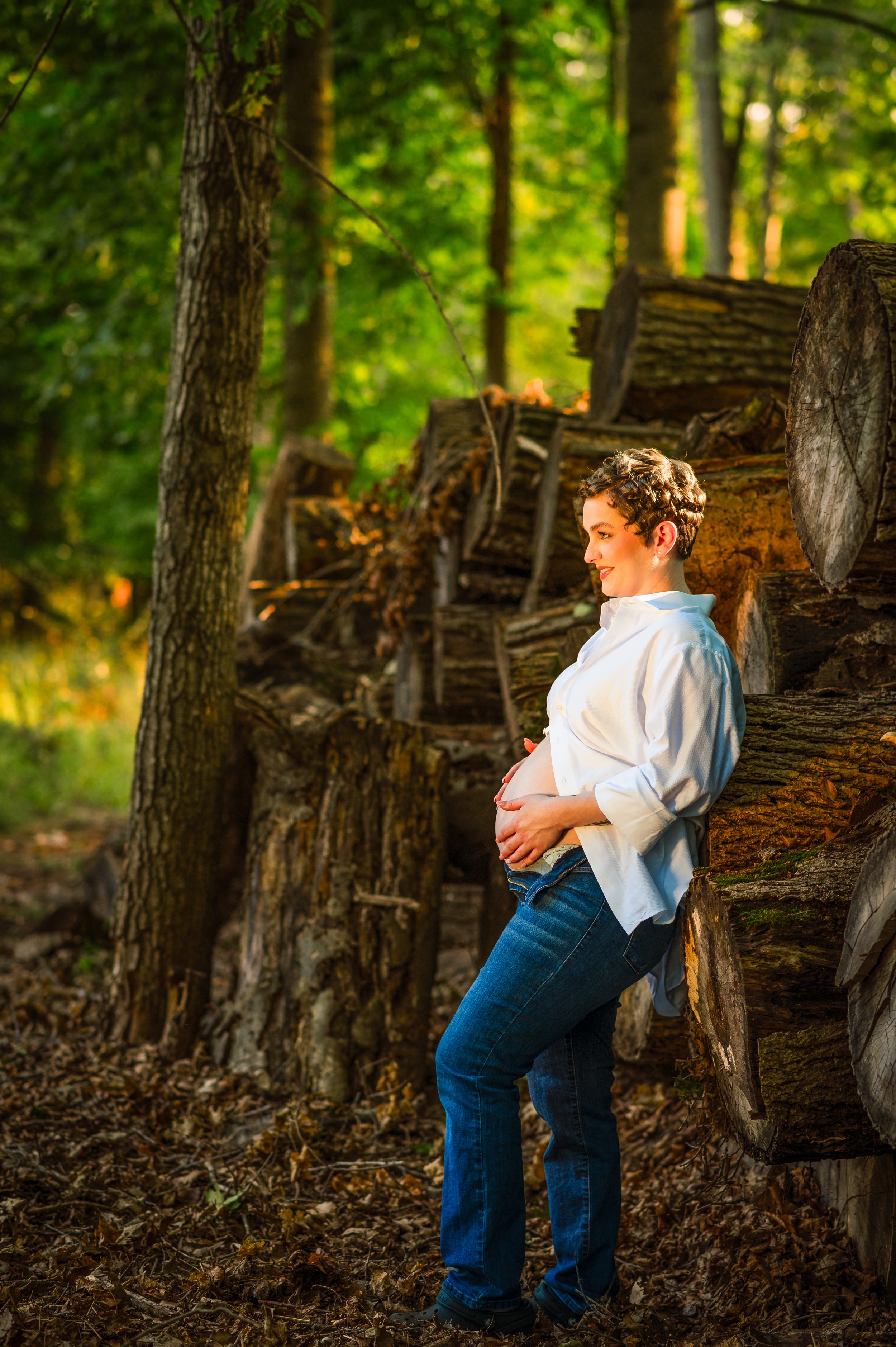 Pregnant woman with short curly hair leaning against a pile of cut logs in a forest, wearing a white shirt and blue jeans, smiling gently.