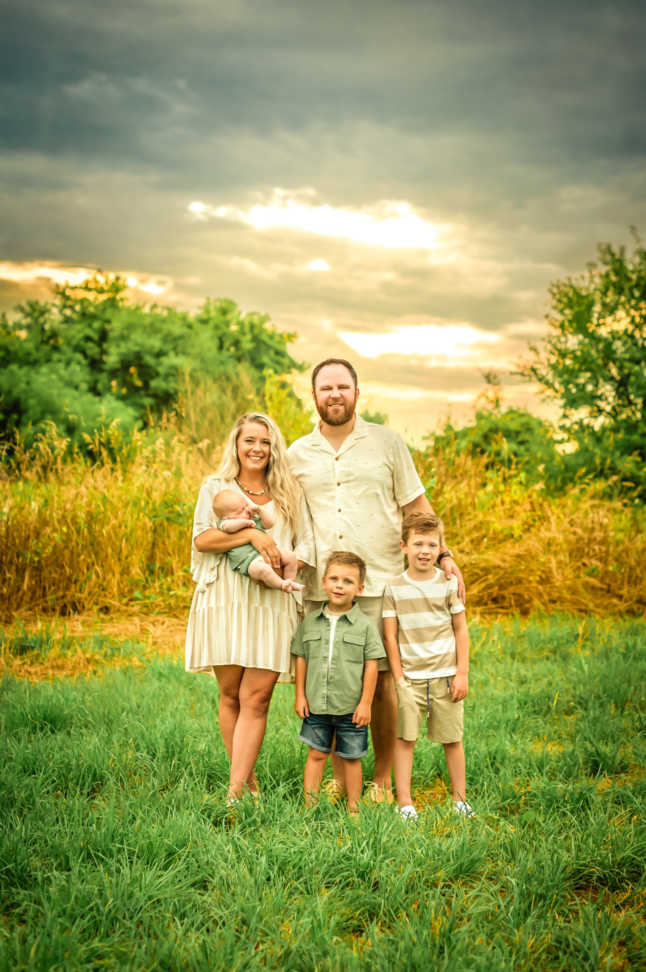 A family of five standing in a grassy field during sunset, with a cloudy sky and trees in the background.