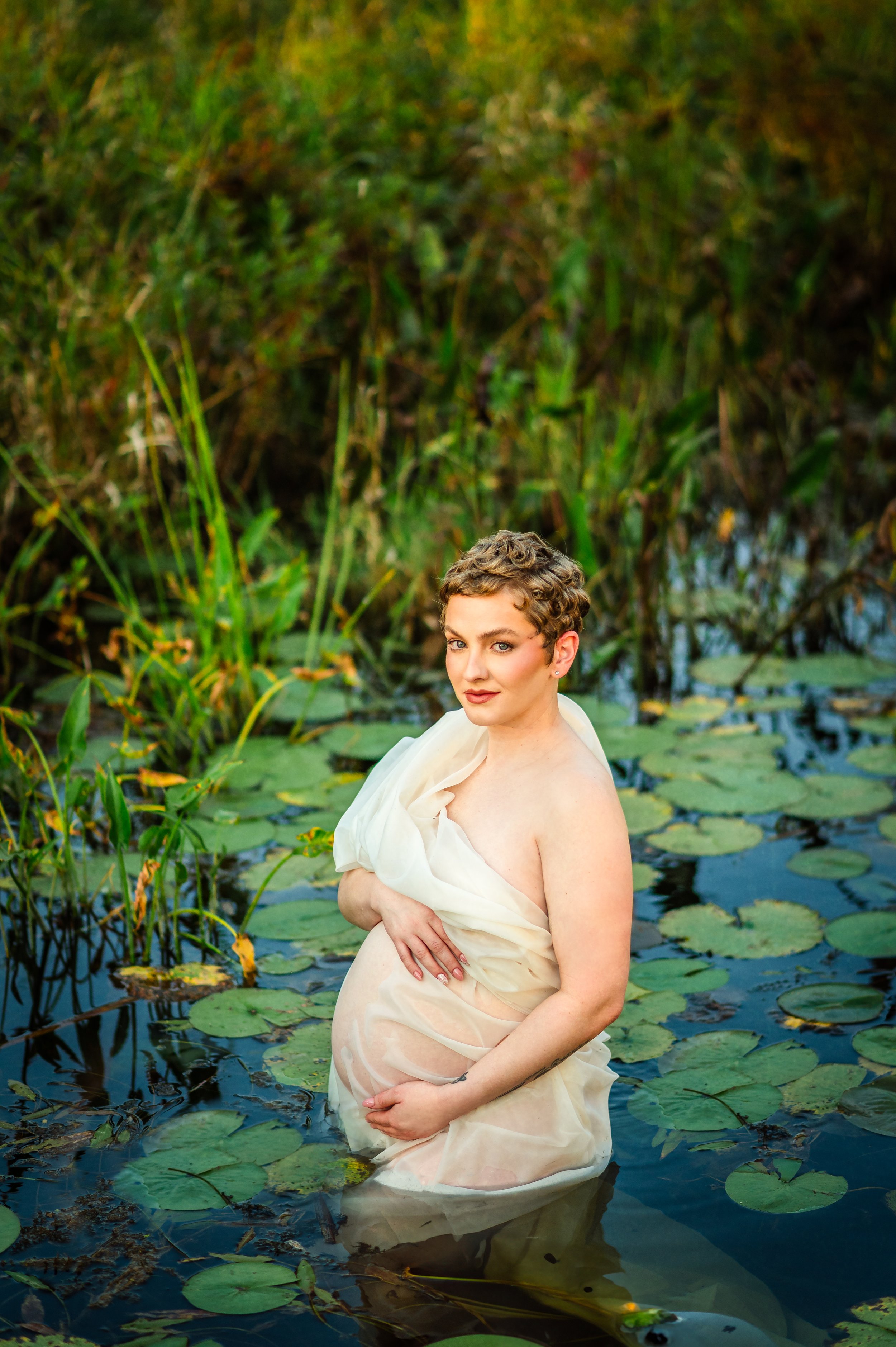 A pregnant woman with short curly hair standing in a pond with lily pads, holding her baby bump and wearing a sheer, off-white dress solo outdoors.