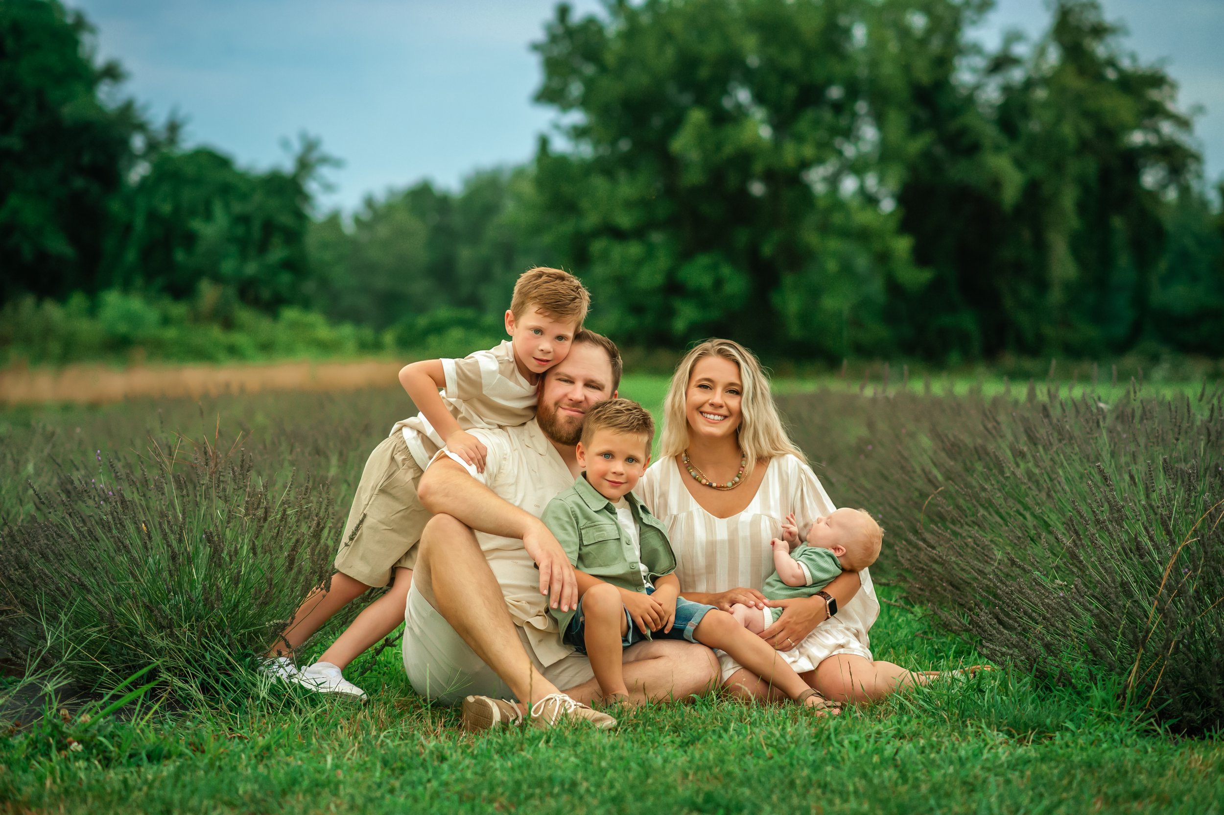 A family of five sitting on grass in a lavender field with trees in the background, smiling at the camera.