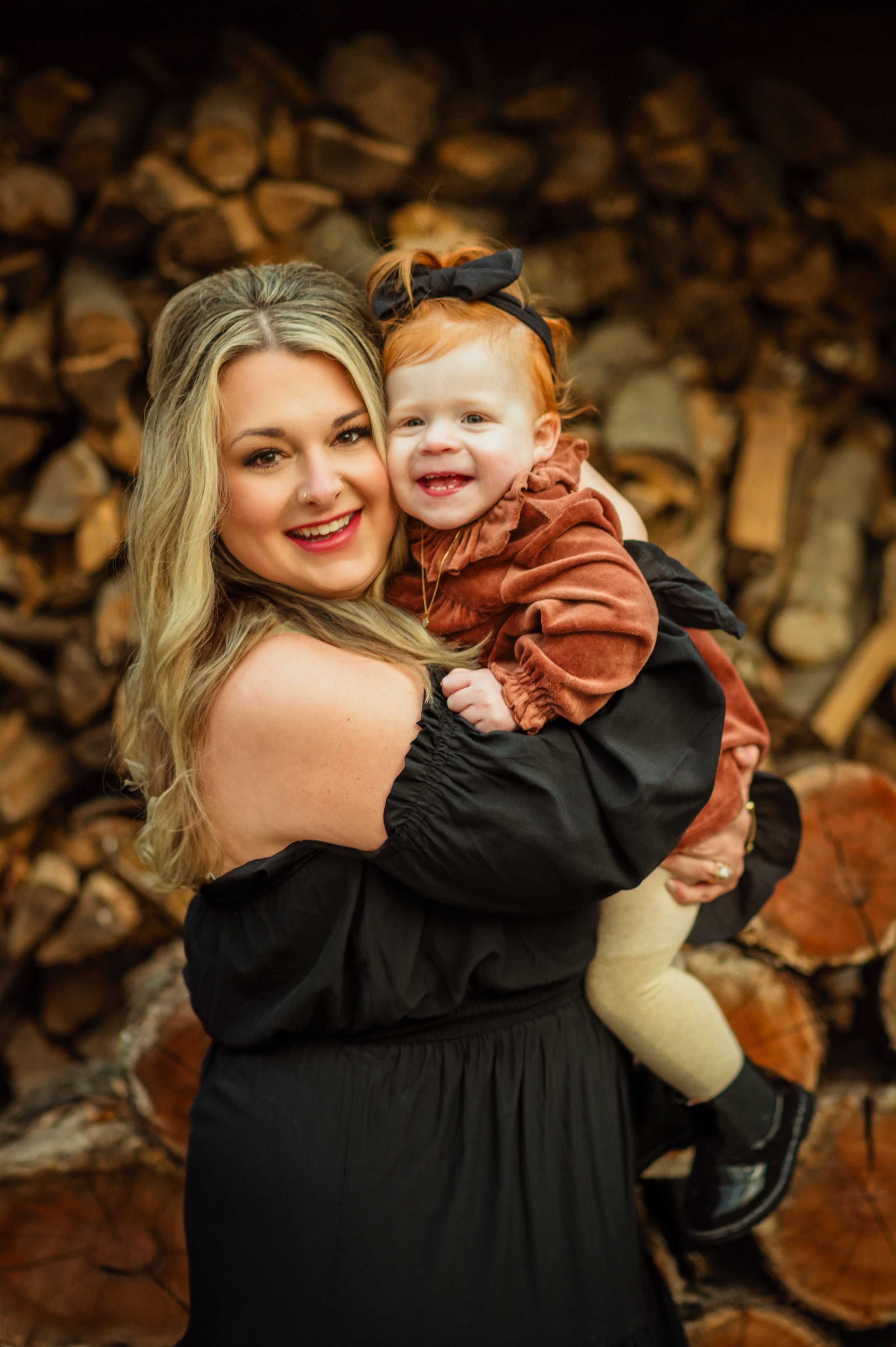 A smiling woman with long blonde hair holding a cheerful young girl with red hair and a black bow in her hair, against a backdrop of stacked firewood.