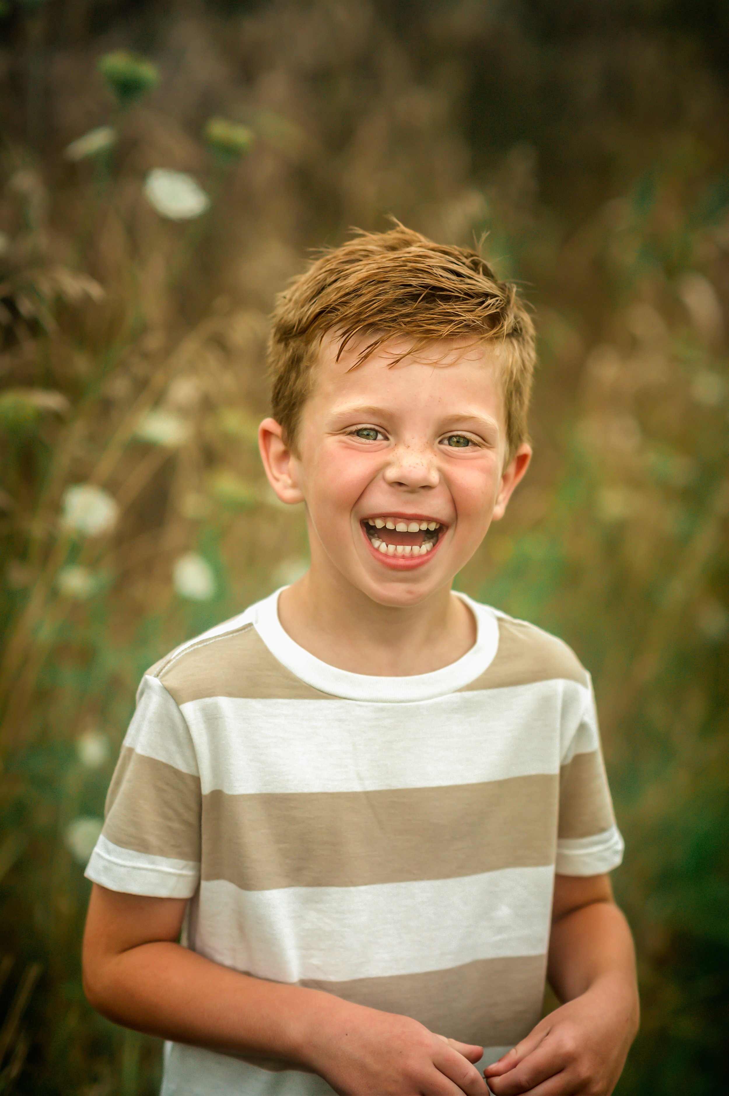 Smiling young boy with short, light brown hair, wearing a beige and white striped t-shirt, standing outdoors with greenery in the background.
