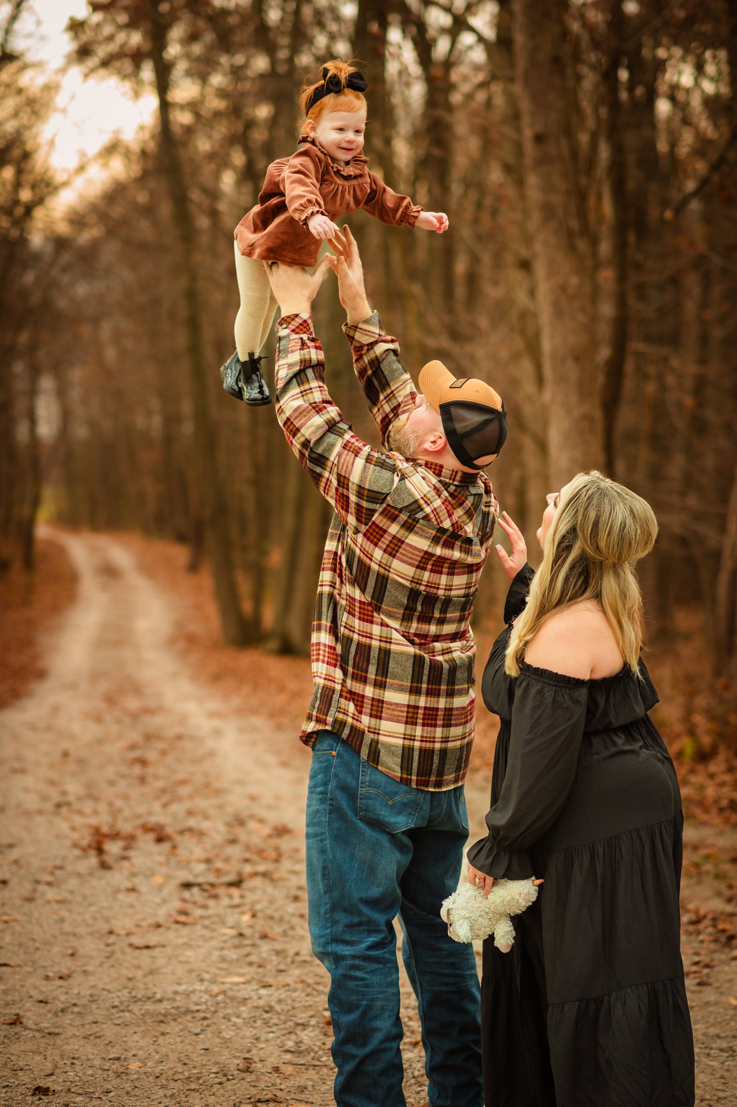 A family with a man, woman, and young girl enjoying an outdoor walk in a forested area. The man lifts the girl into the air while the woman smiles and looks on.