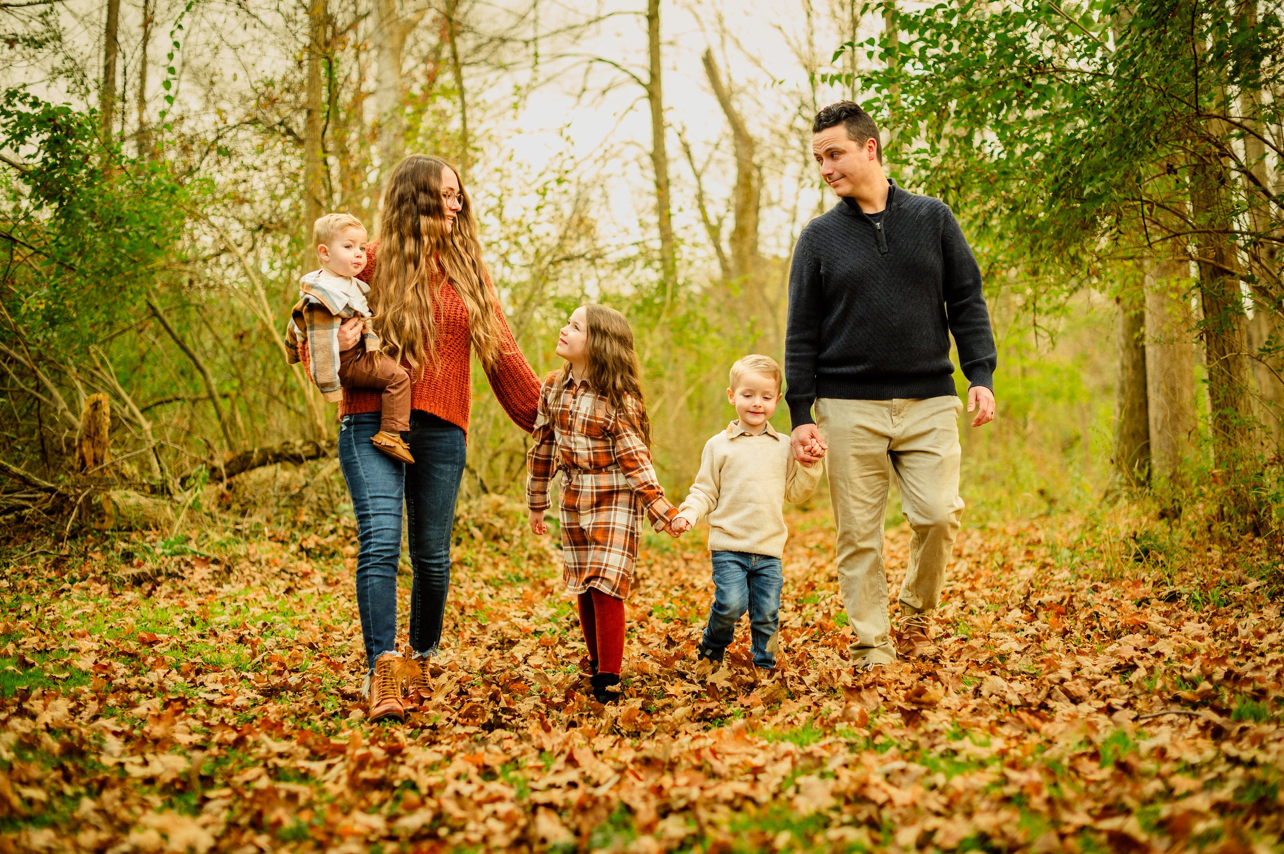 A family of six walking in a wooded area during autumn, with fallen leaves on the ground. The mother is holding a young child, and the father is holding the hand of a young boy. Two girls are walking holding hands, with the family enjoying a walk in