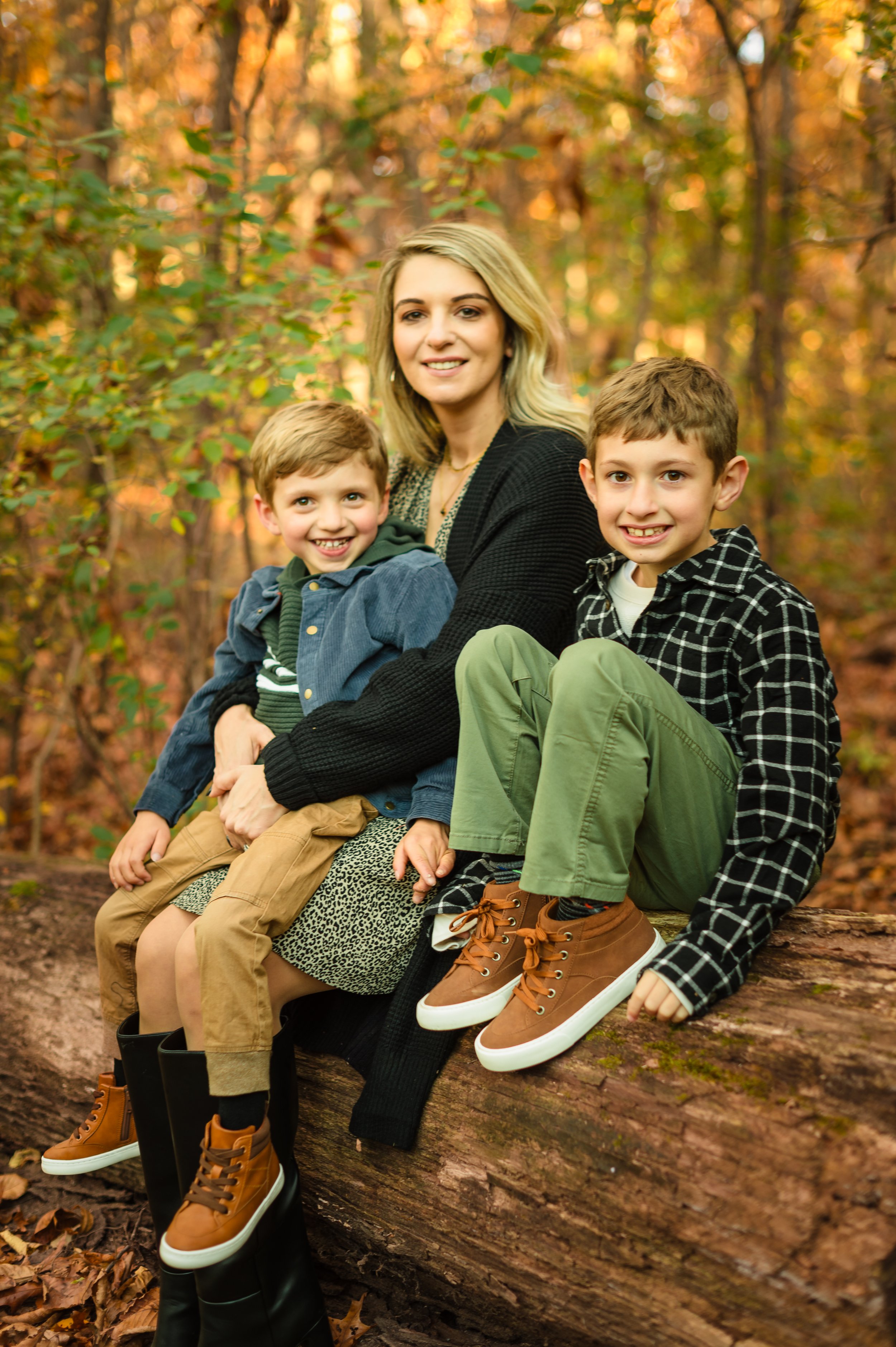 A woman and two young boys sitting on a fallen log in a forest during autumn, smiling at the camera.